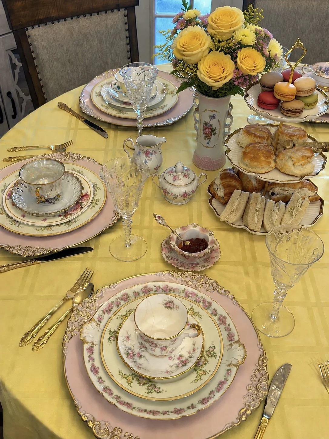 A fancy table setting with floral china, gold silverware, wine glasses, a bouquet of yellow and pink roses in a porcelain vase, and a tray of assorted macarons, pastries, and sandwiches on a yellow tablecloth.