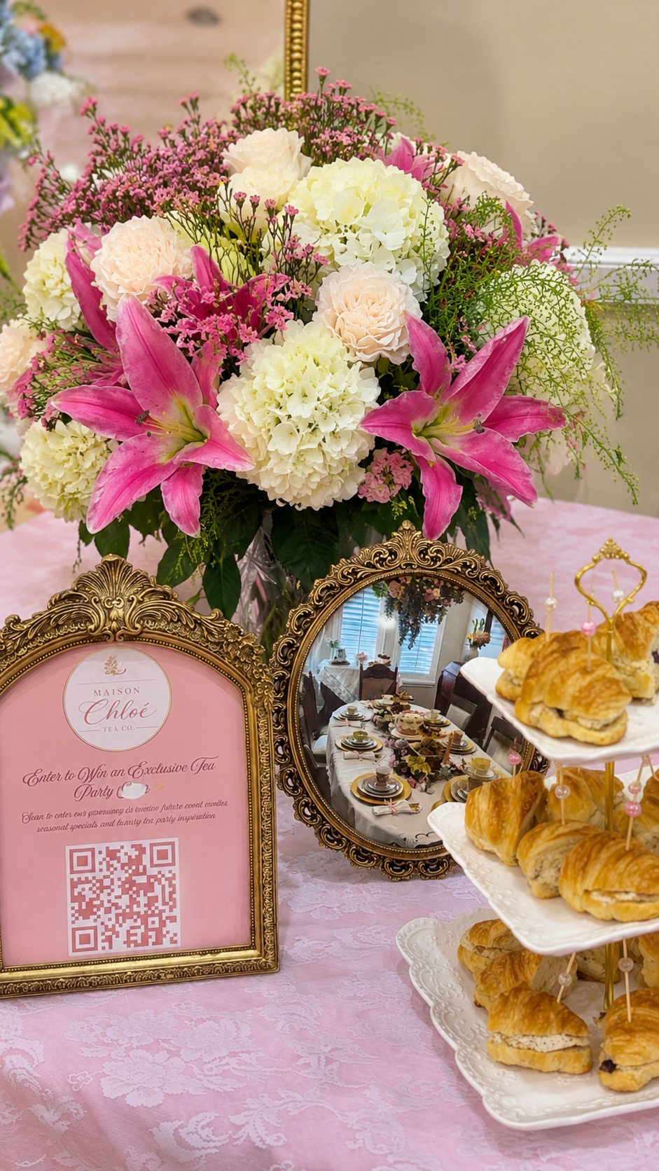 Elegant table setup with a large bouquet of pink and white flowers, a framed mirror, a tiered plate of croissants, and a pink sign advertising an exclusive tea party with a QR code, all arranged on a pink tablecloth.