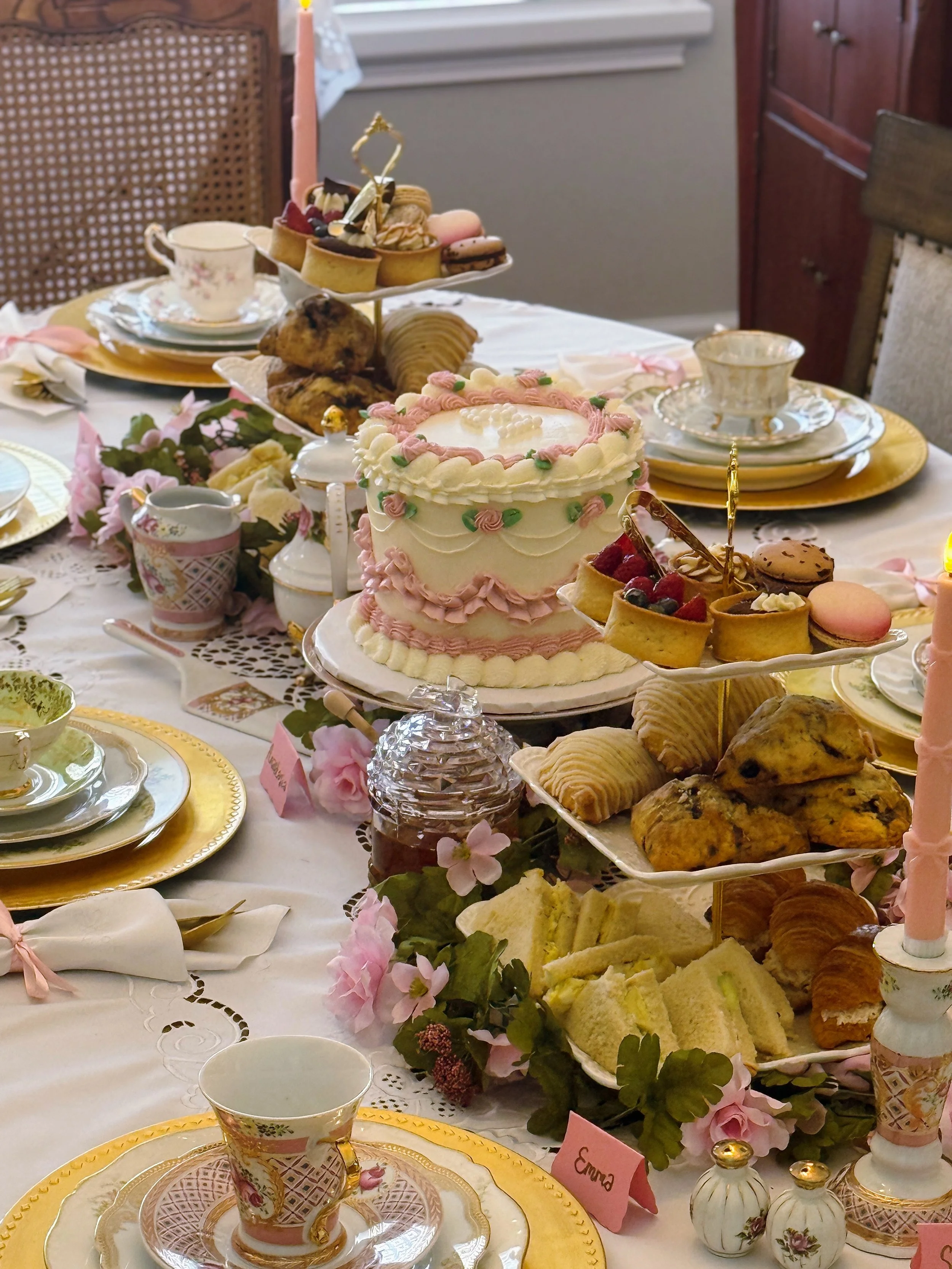 A table set for a celebration with a variety of desserts, including cake, cookies, and pastries, decorated with pink flowers and elegant china dishes with gold accents.