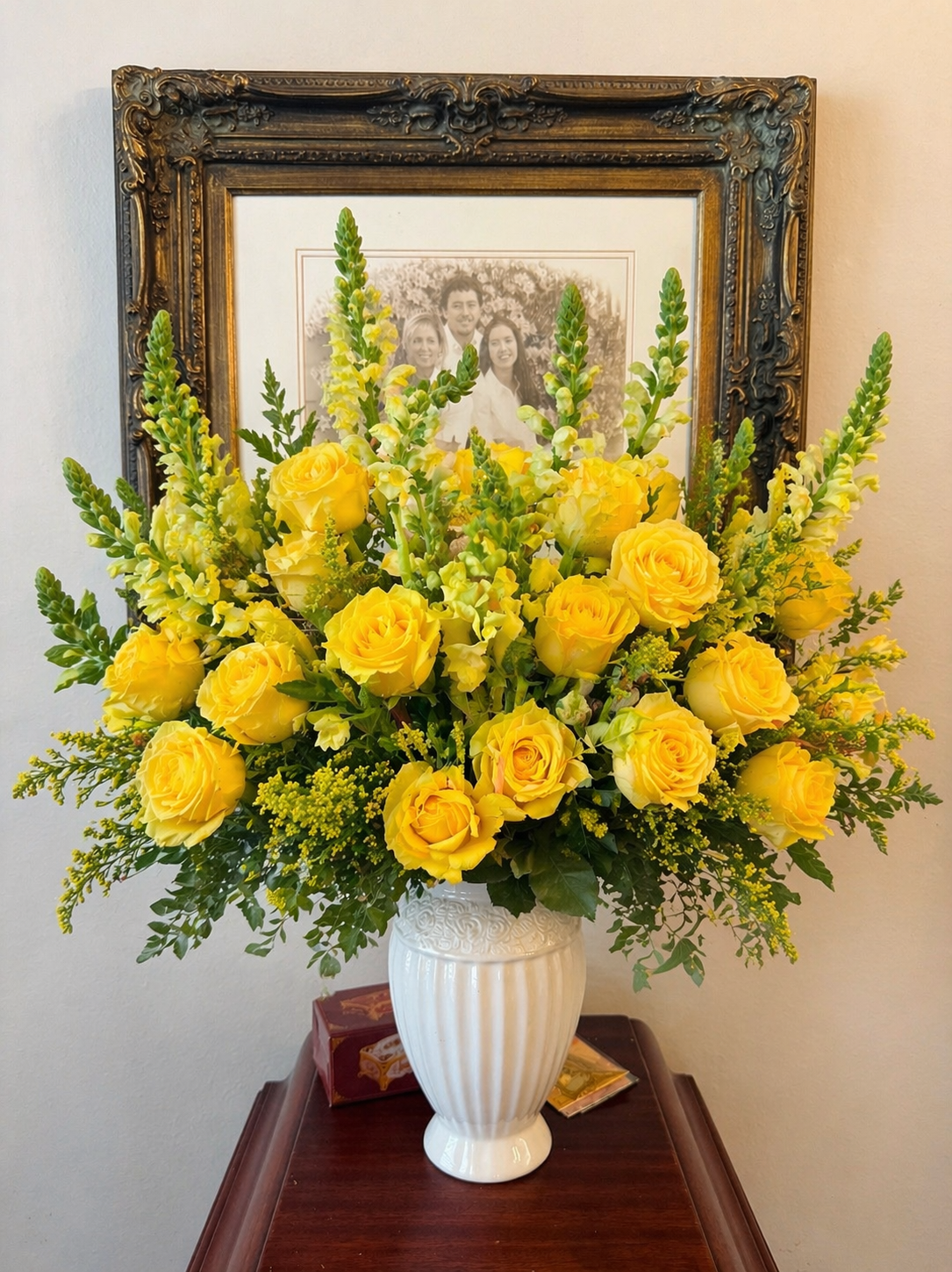 Yellow flower arrangement with roses and other flowers in a white vase, placed on a wooden table in front of a framed family photo.