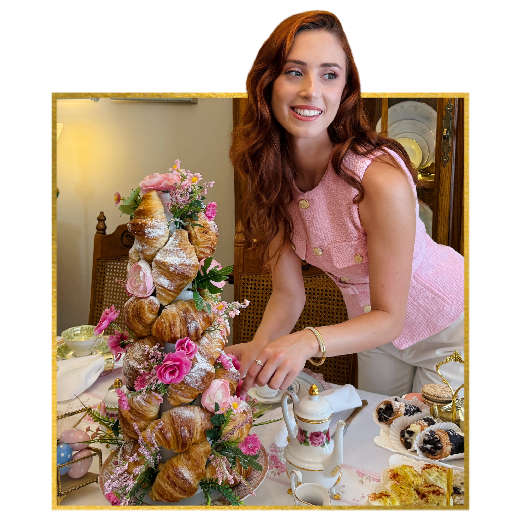 A woman with red hair smiling at a birthday party decorated with flowers and a large stack of croissants, assorted desserts, and desserts on the table.