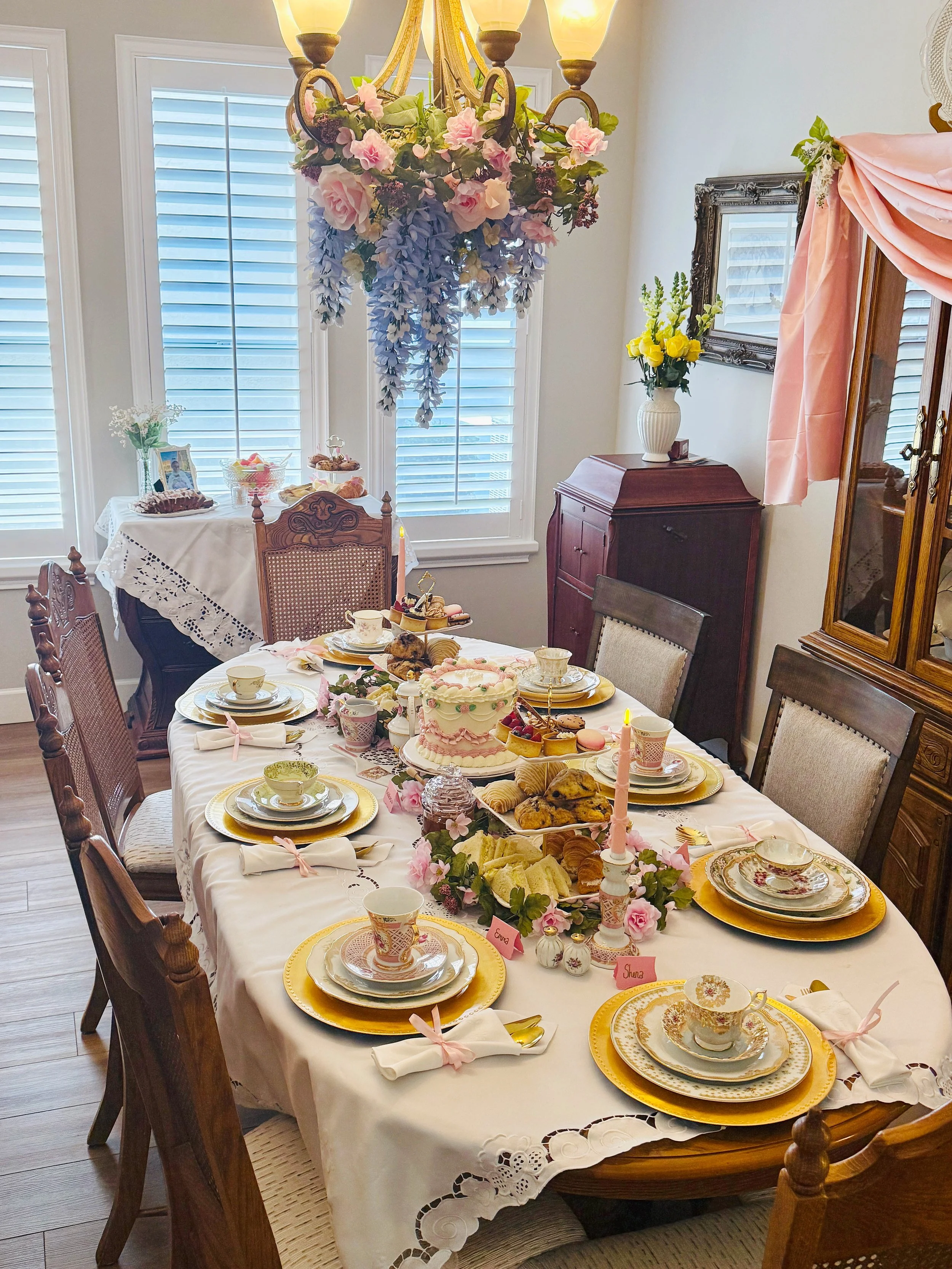 A dining table set for a celebration, possibly a birthday or bridal shower, with a white lace tablecloth, gold-rimmed fine china, teacups, and plates, decorated with pink and white flowers and pink ribbons. The table has various desserts including ti