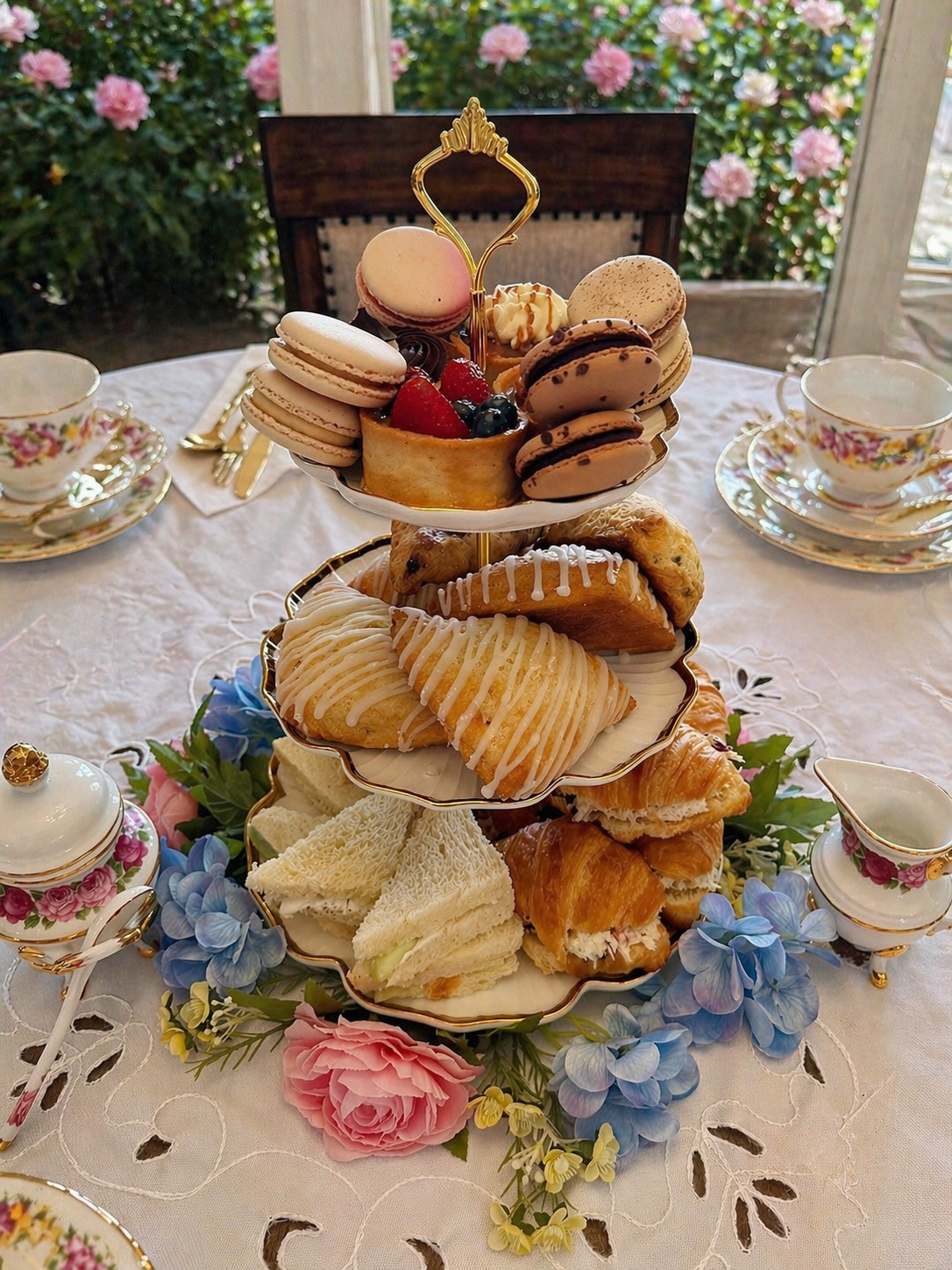 A multi-tiered tray of assorted desserts including macarons, fruit tart, pastries, and scones, surrounded by floral decorations on a dining table set with fine china.