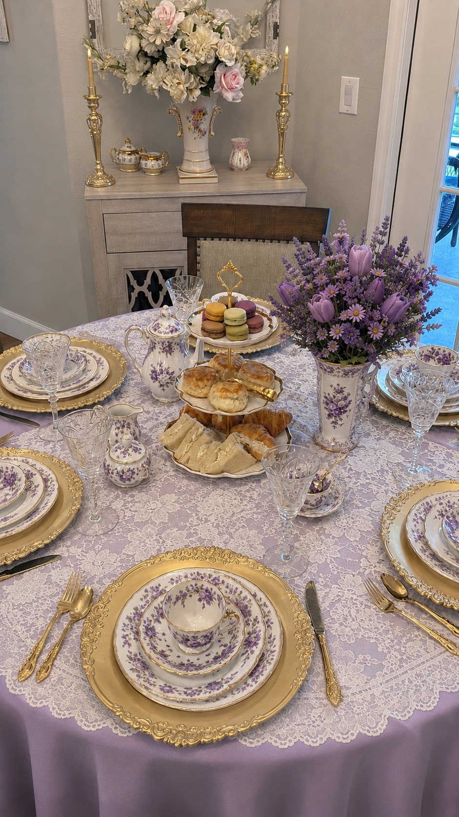 A beautifully set dining table with purple and gold floral china, gold flatware, and crystal glasses. The table has a lavender tablecloth and a lace overlay. There is a vase of purple flowers and a cake stand with macarons, pastries, and sandwiches. 