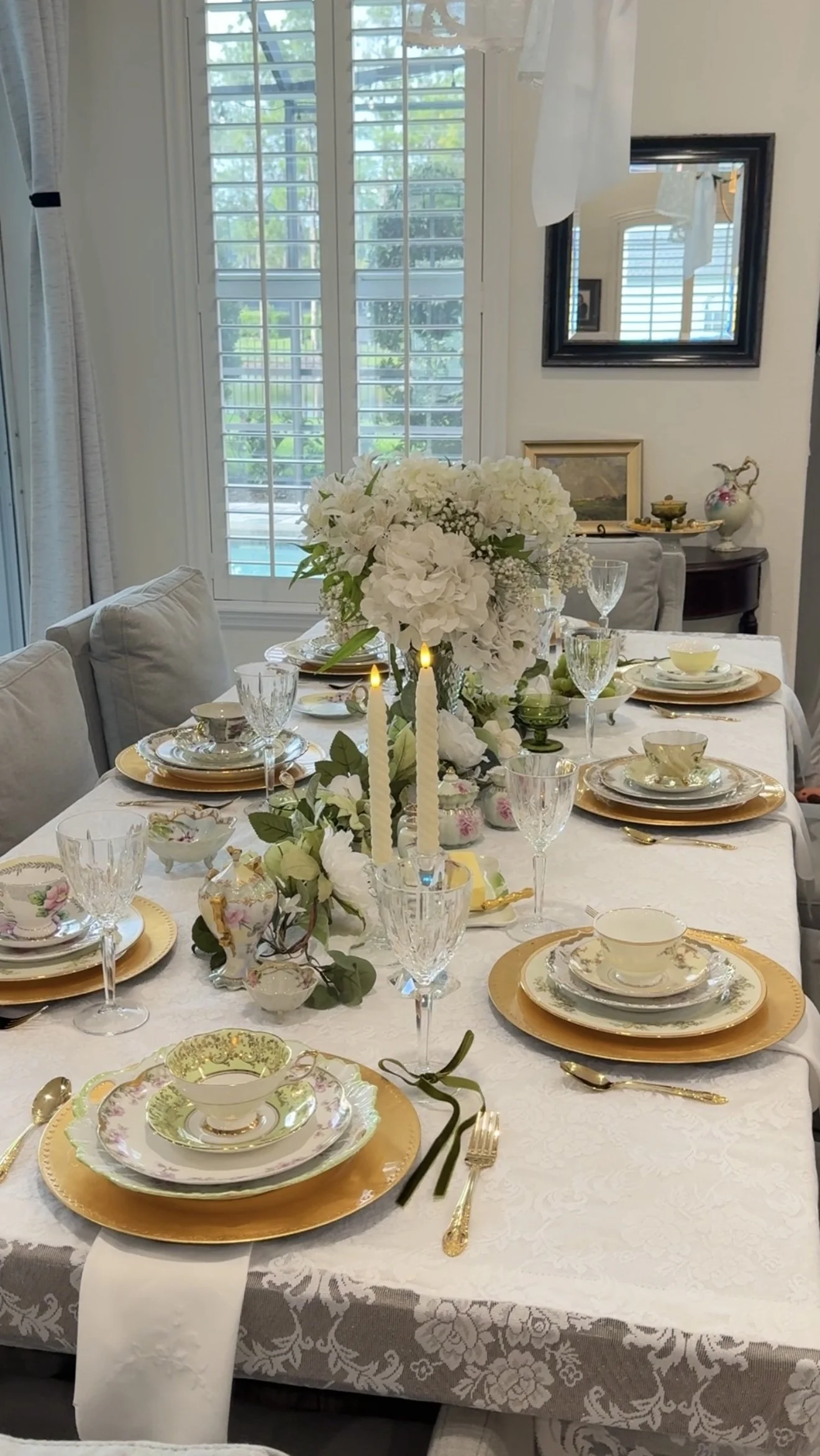 Elegant dining table set with fine china, gold accents, wine glasses, and floral centerpiece, in a bright room with large windows and white shutters.