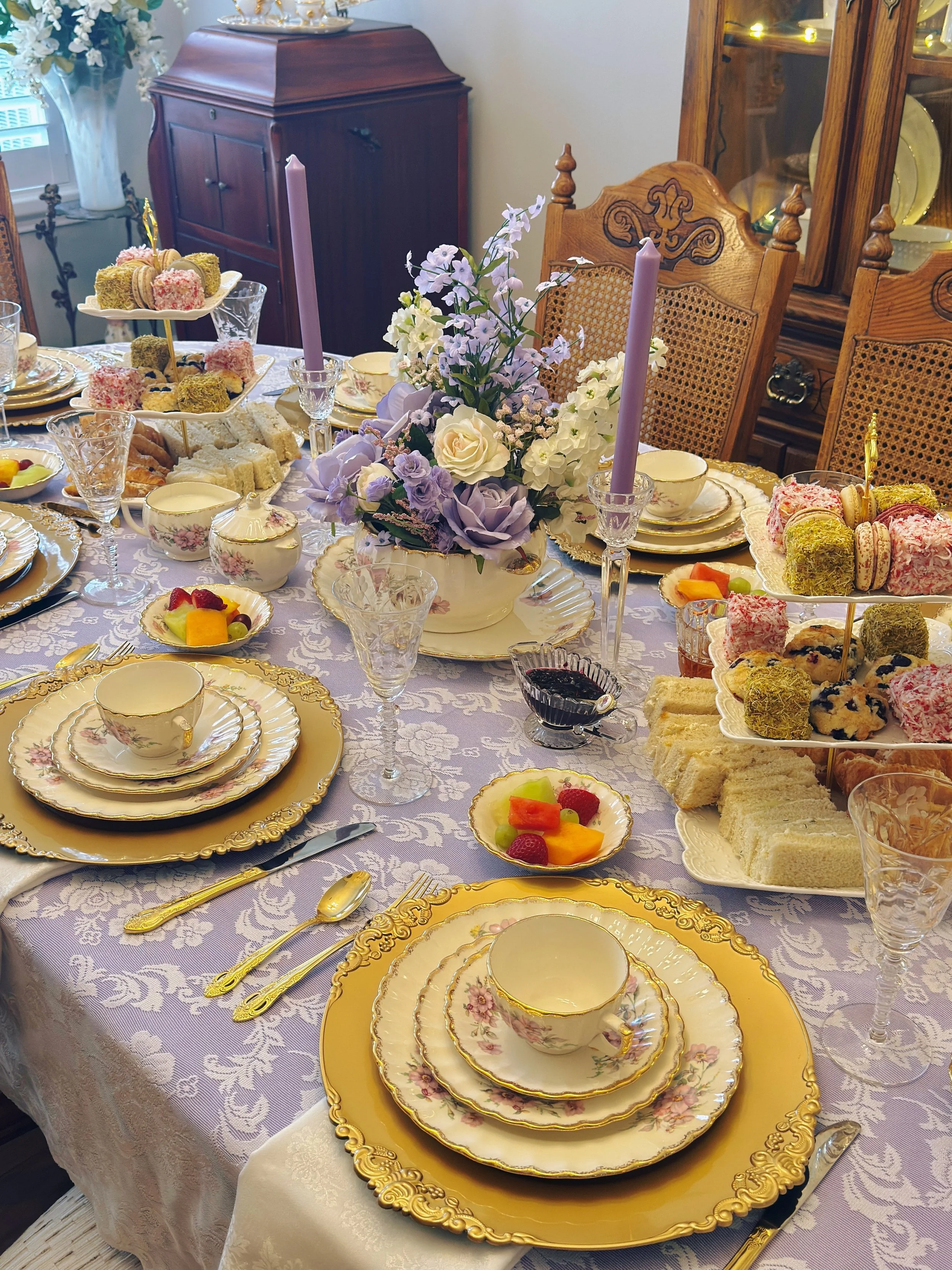 A formal dining table set with fine china, gold-rimmed plates, cups, and saucers, along with gold cutlery. The table features a centerpiece with purple, white, and green flowers and tall pastel purple candles. There are various desserts, including fr