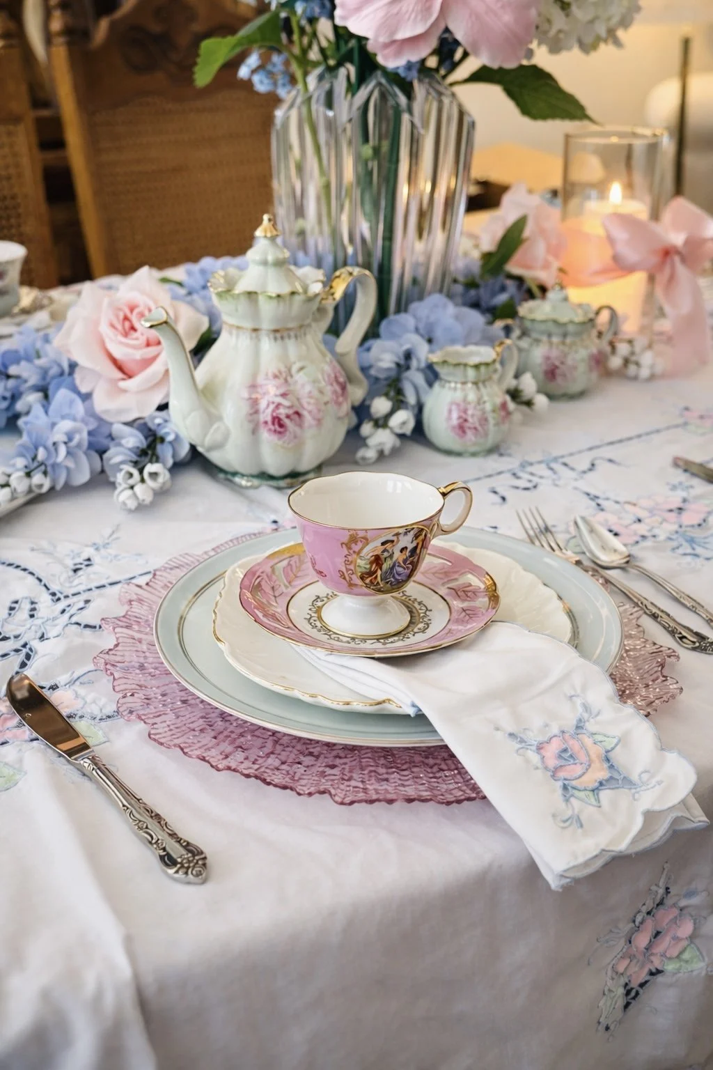 An elegant table setting with floral china, pink and gold accents, and a centerpiece of pink and white roses and light blue hydrangeas in a glass vase. There are teapots, sugar bowls, a candle, and a pink ribbon decor on the table.