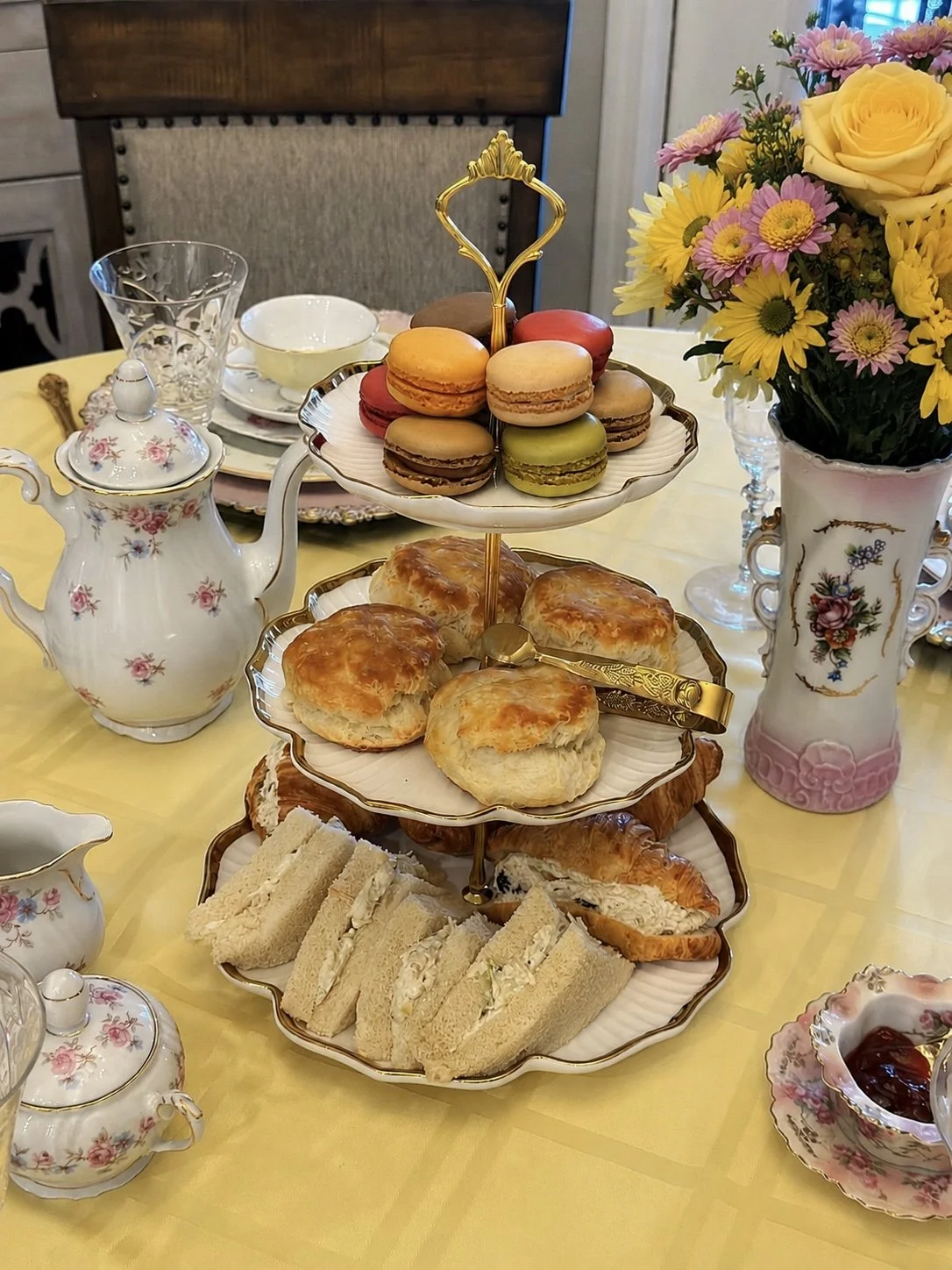 A tea table with a floral teapot, a tiered tray with assorted macarons and scones, slices of finger sandwiches, a vase of pink and yellow flowers, and fine china dishes.