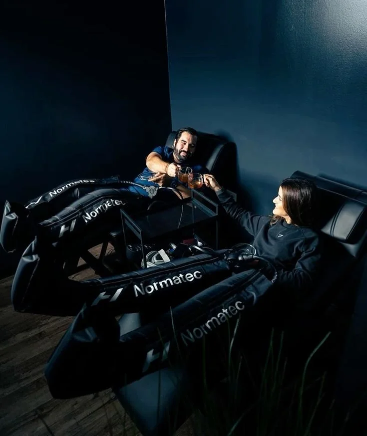 Two people sitting in Normatec compression boots, toasting with glasses of water in a dark room.