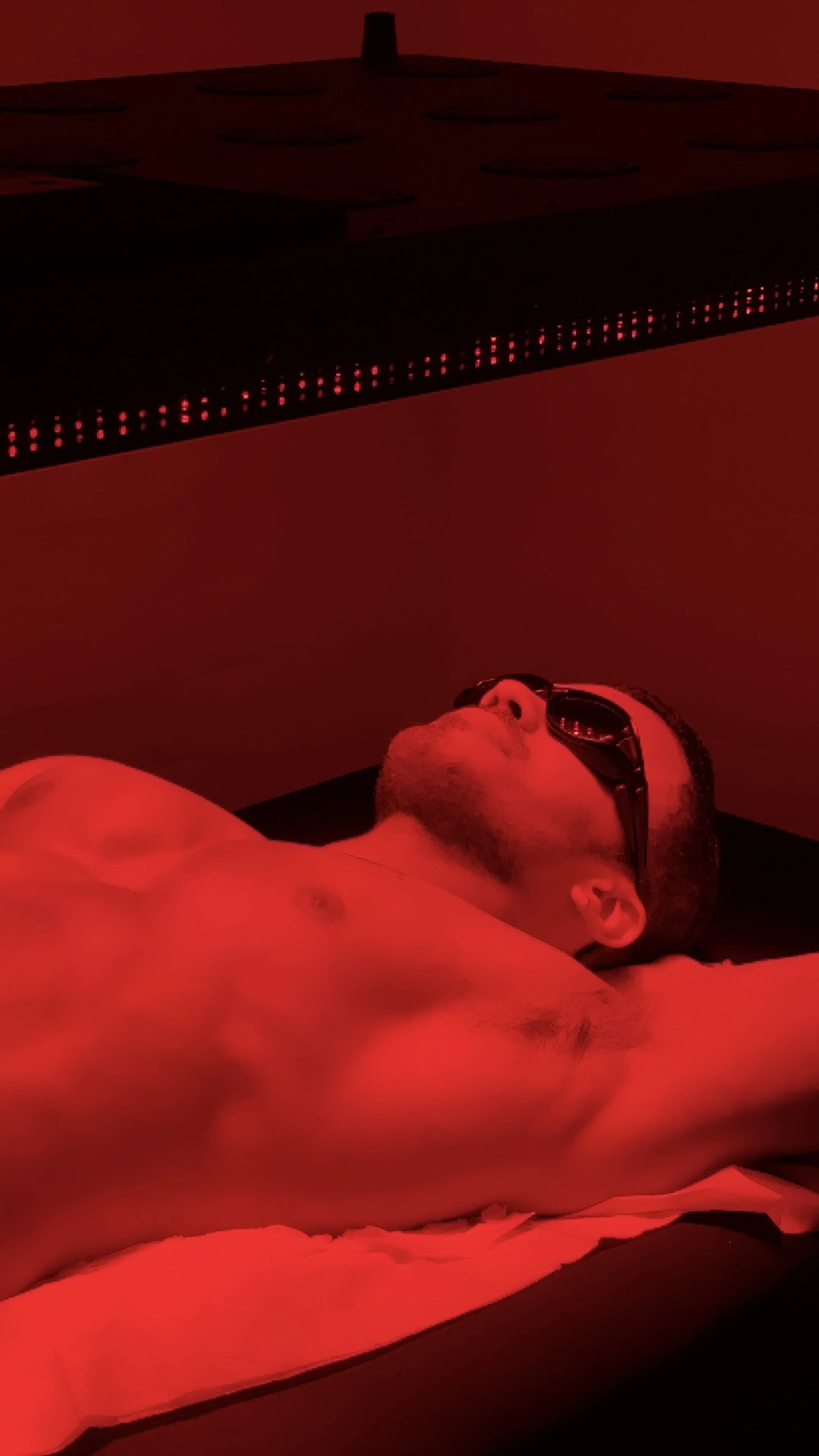 A person with dark hair and sunglasses lying on a treatment bed under a red light therapy panel.