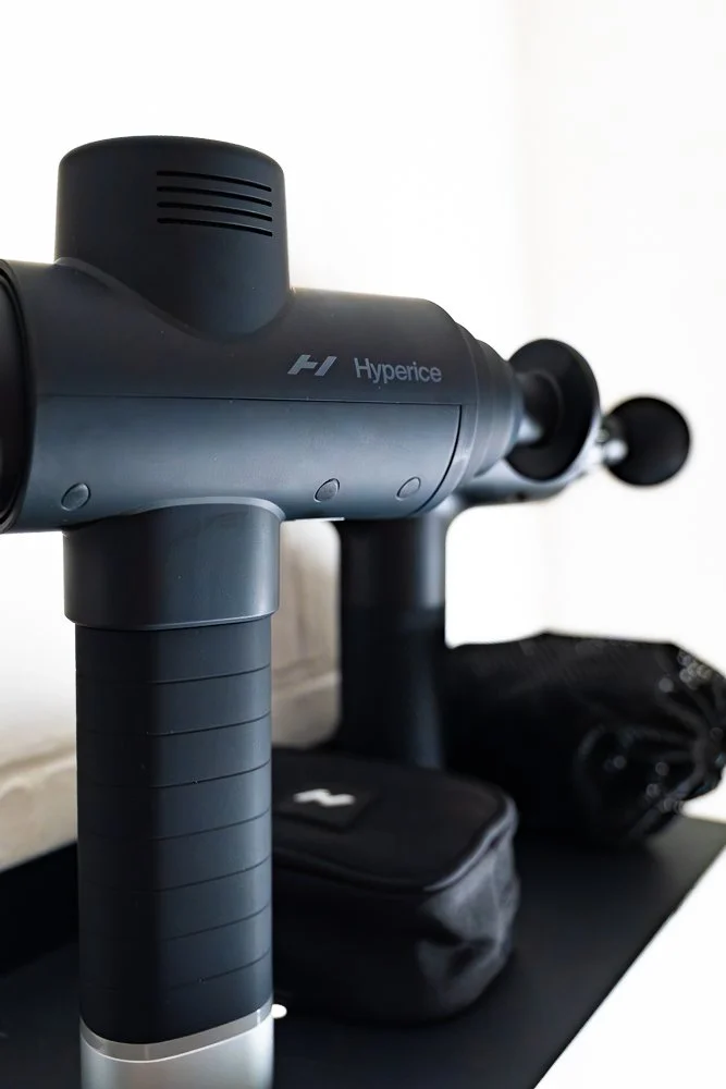 A Hyperice massage gun placed on a black surface, with a black carrying case behind it and a black pouch to the right.