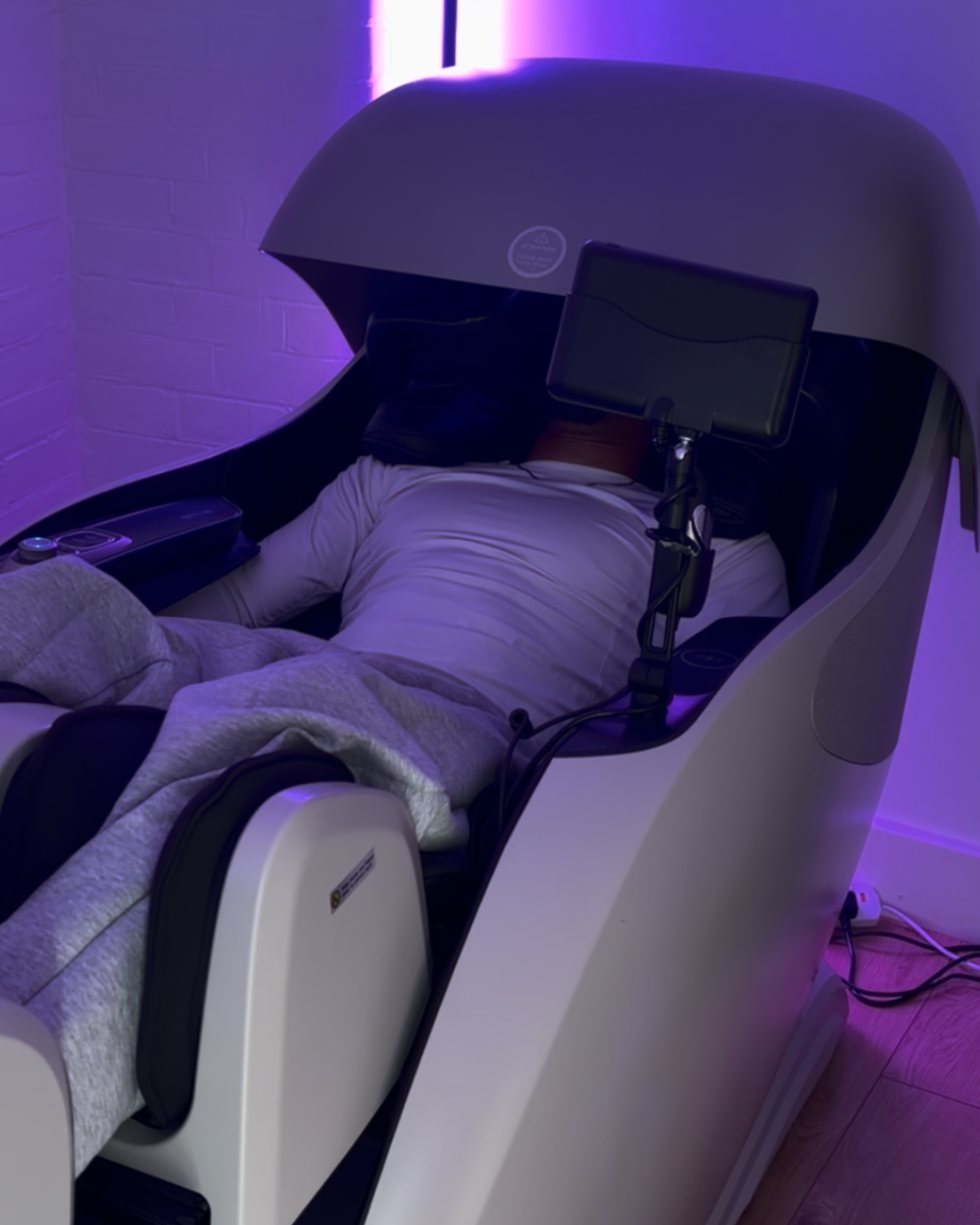 Person lying inside a futuristic massage machine with a privacy hood, surrounded by purple ambient lighting.