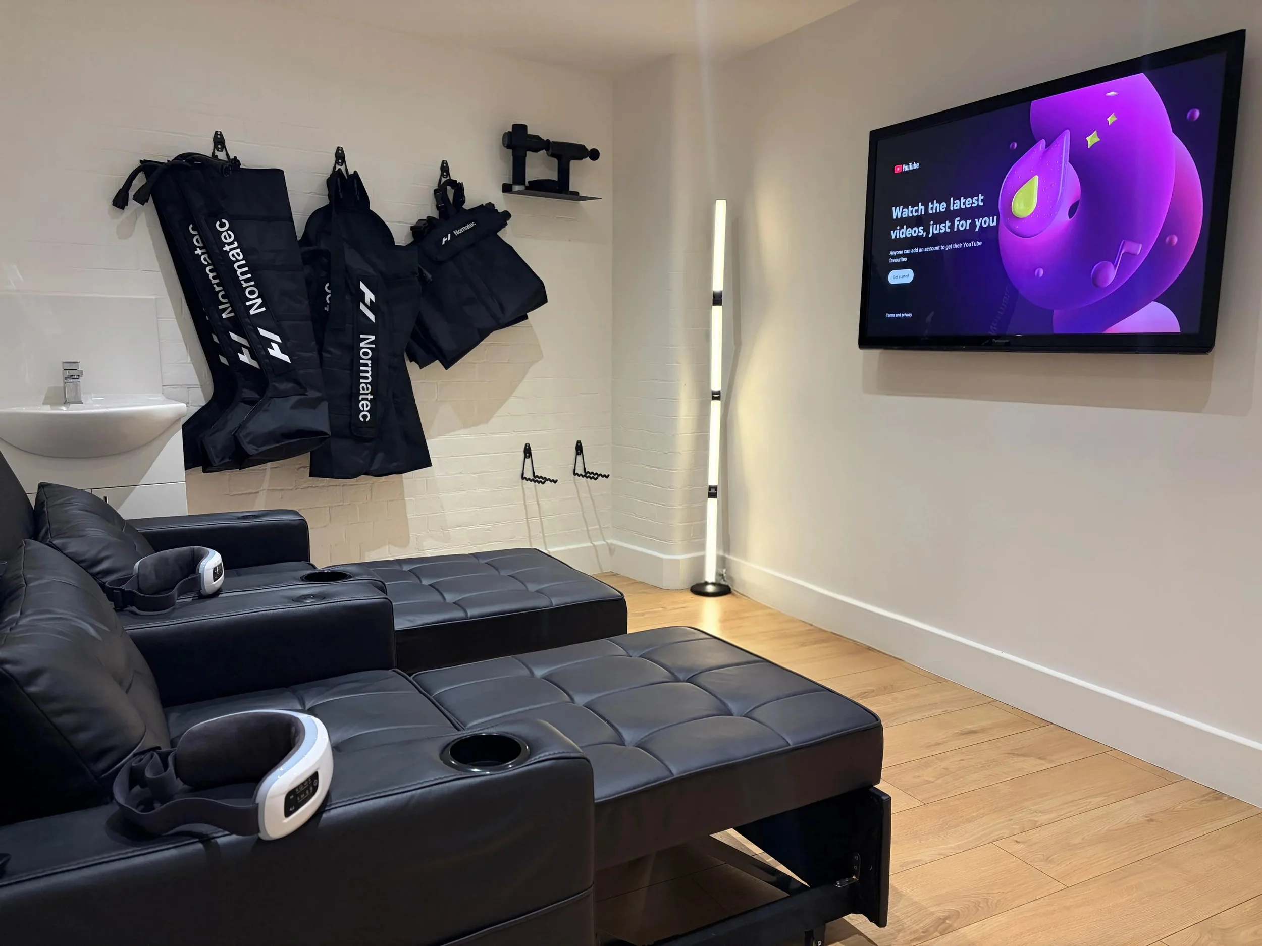 Sports clinic space with a black leather sectional sofa, a TV mounted on the wall displaying YouTube, and Normatec and Hyperice recovery equipment hanging on hooks. There is a white brick wall,  a vertical LED light, and wood flooring.