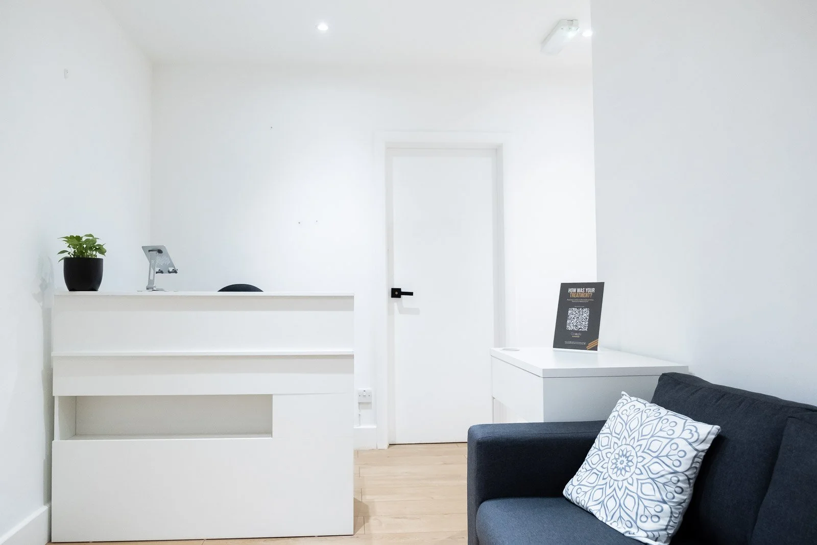 Minimalist office reception area with white walls, a white counter with a small potted plant, a black chair behind the counter, a white door in the background, and a black sofa with white and blue patterned pillows. There is a side table with a sign and QR code.