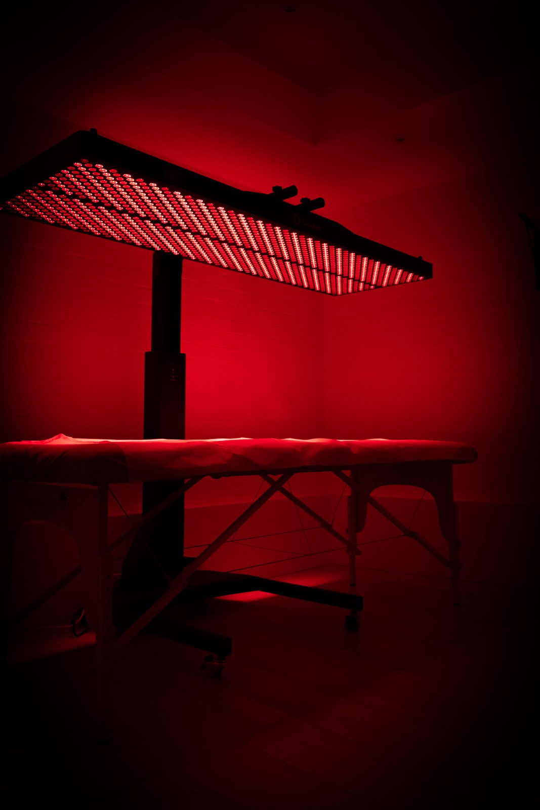 Long red LED therapy panel over a massage table in a dark room.