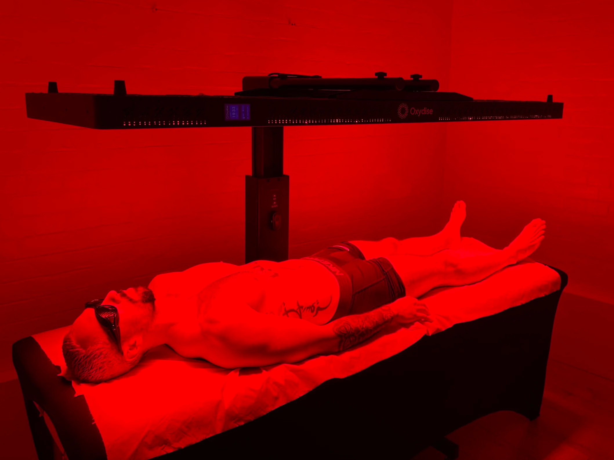 Person lying on a bed in a red light therapy room with equipment labeled 'Oxydise' above.
