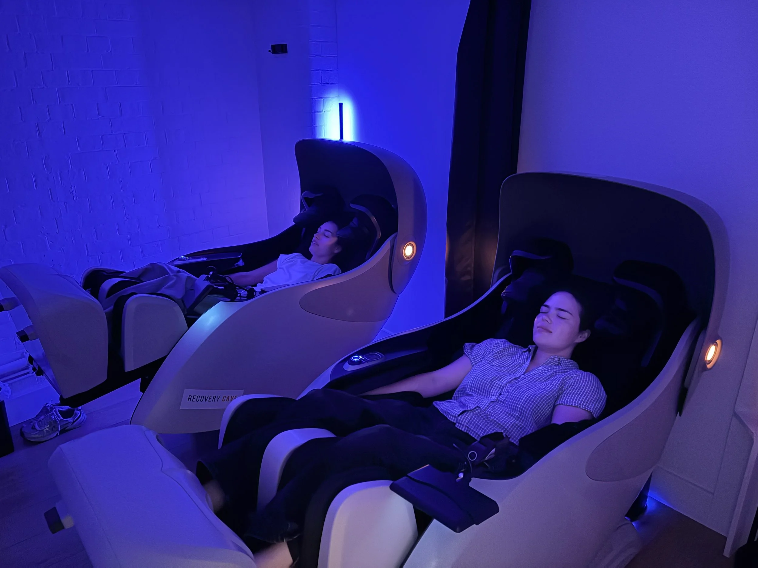 Two women lying in futuristic massage machines in a sensory room with soft purple lighting, they are dressed casually and are relaxing with their eyes closed