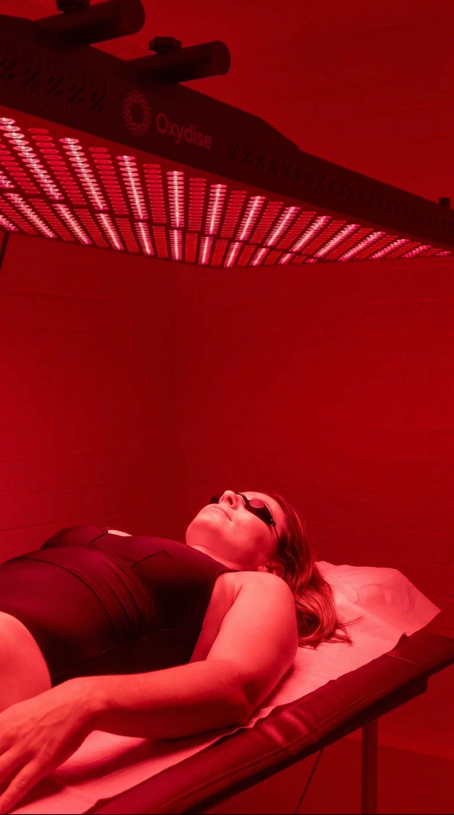 A person lying on a bed under red LED light therapy, wearing protective glasses.