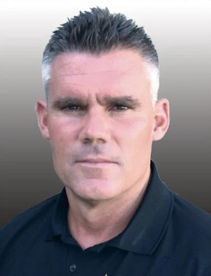 A man with short, dark hair and a serious expression wearing a black collared shirt.