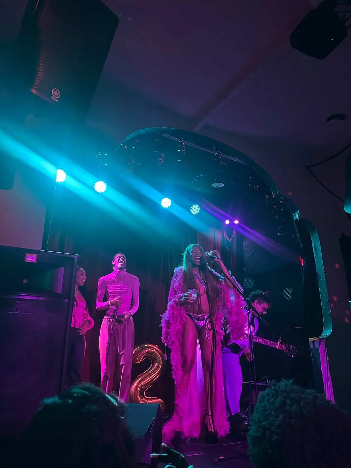 A singer stands on stage with musicians under colorful pink and blue stage lights. The singer is wearing a pink feathered outfit and holding a microphone, with two women and one man in the background, all dressed in pink. There is a large balloon number 2 on the stage, indicating a celebration or event.