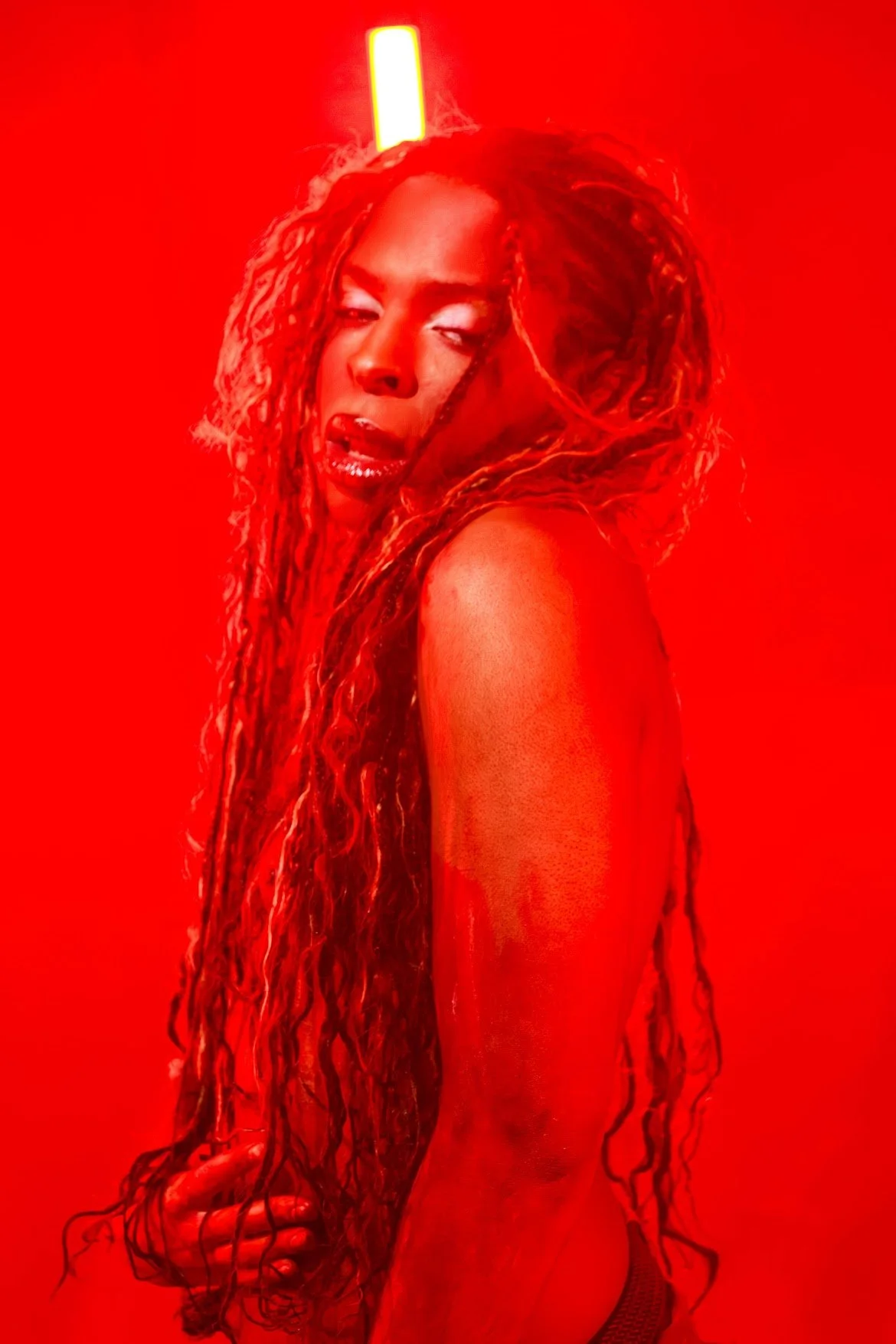 A woman with long, curly hair and shiny makeup posing against a red background with a bright light behind her head.