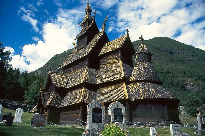 Wooden church with multiple spires and a cross on top, set against a backdrop of green mountains and a partly cloudy sky.