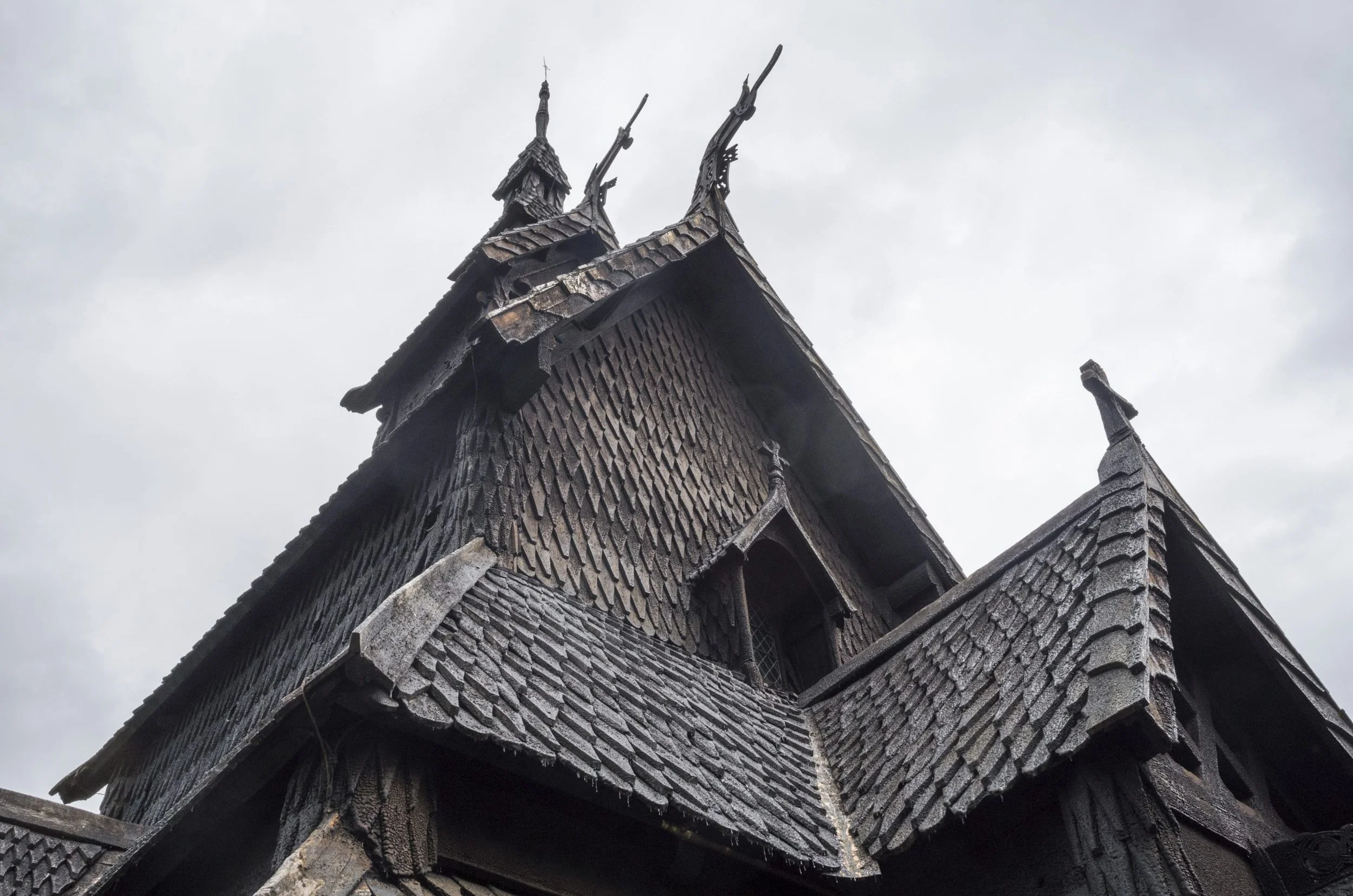 The image shows a close-up of the roof of an old, wooden church with intricate detailing at the gable ends, set against a cloudy sky.