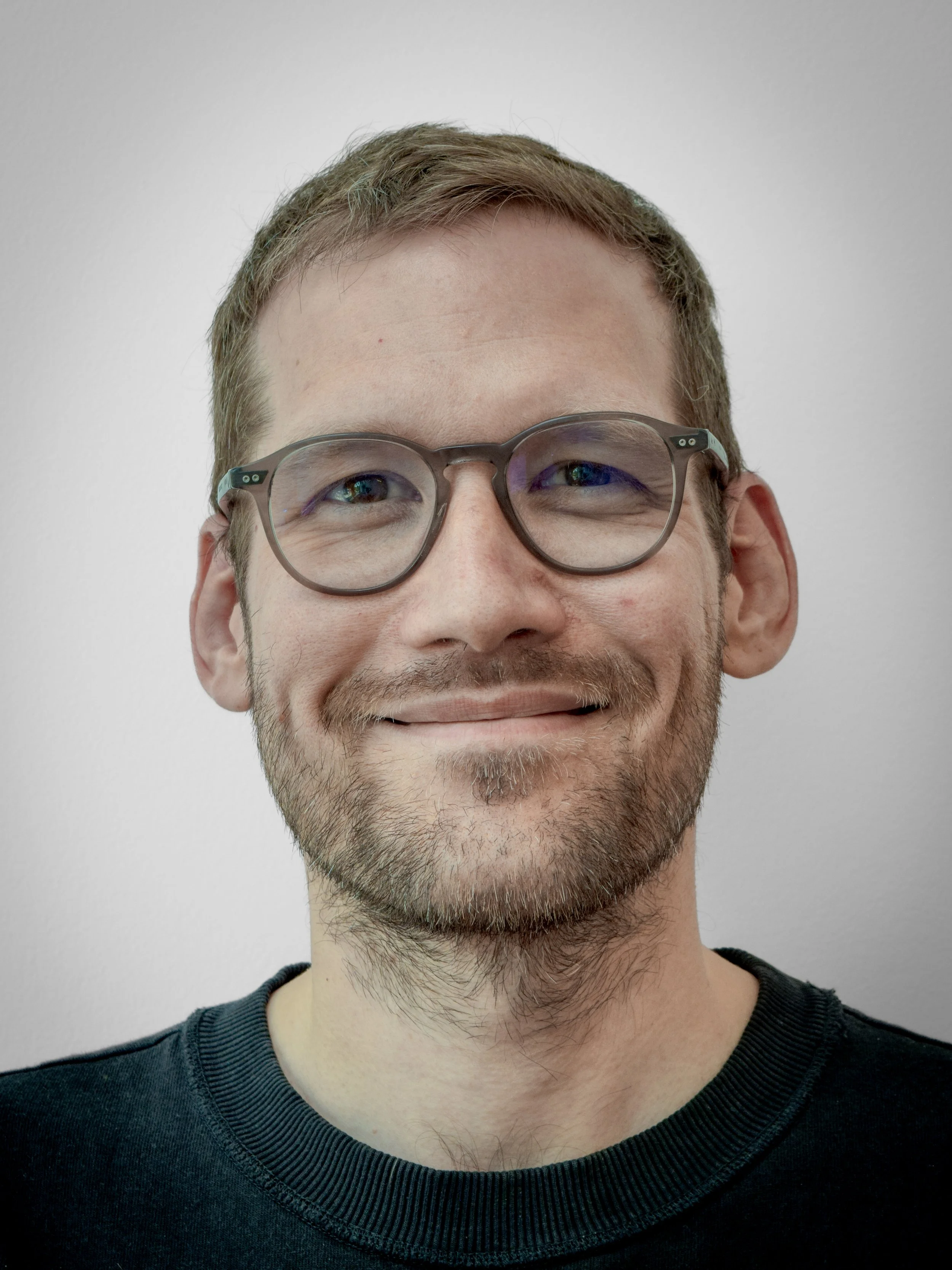Close-up of a smiling man with brown hair, glasses, and a beard, wearing a black shirt, against a plain white background.