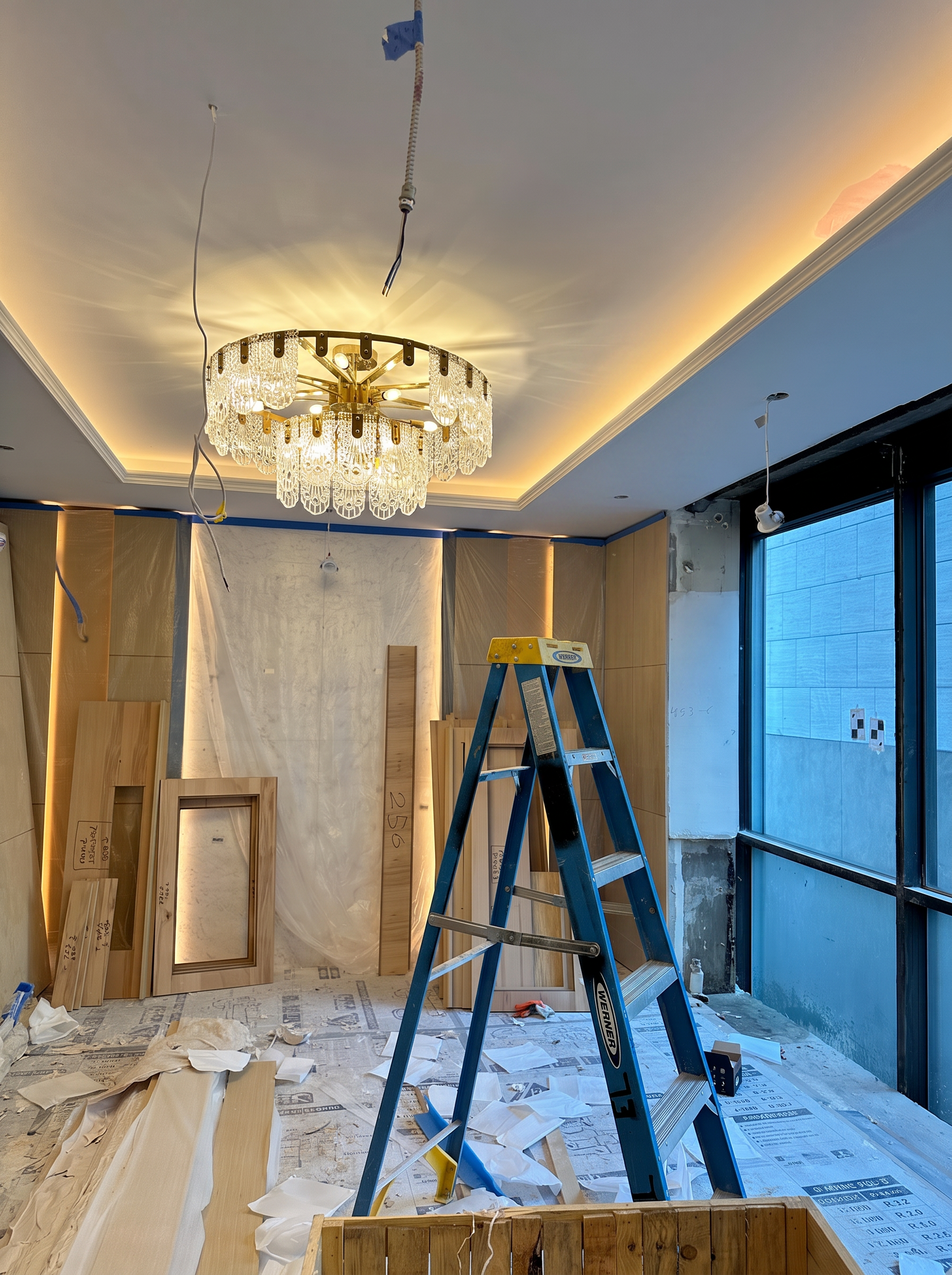 Interior of a room under renovation with cardboard frames, wood planks, a ladder, and construction materials scattered on the floor. Ceiling has partial lighting and wires hanging down, with a large window on the right side.