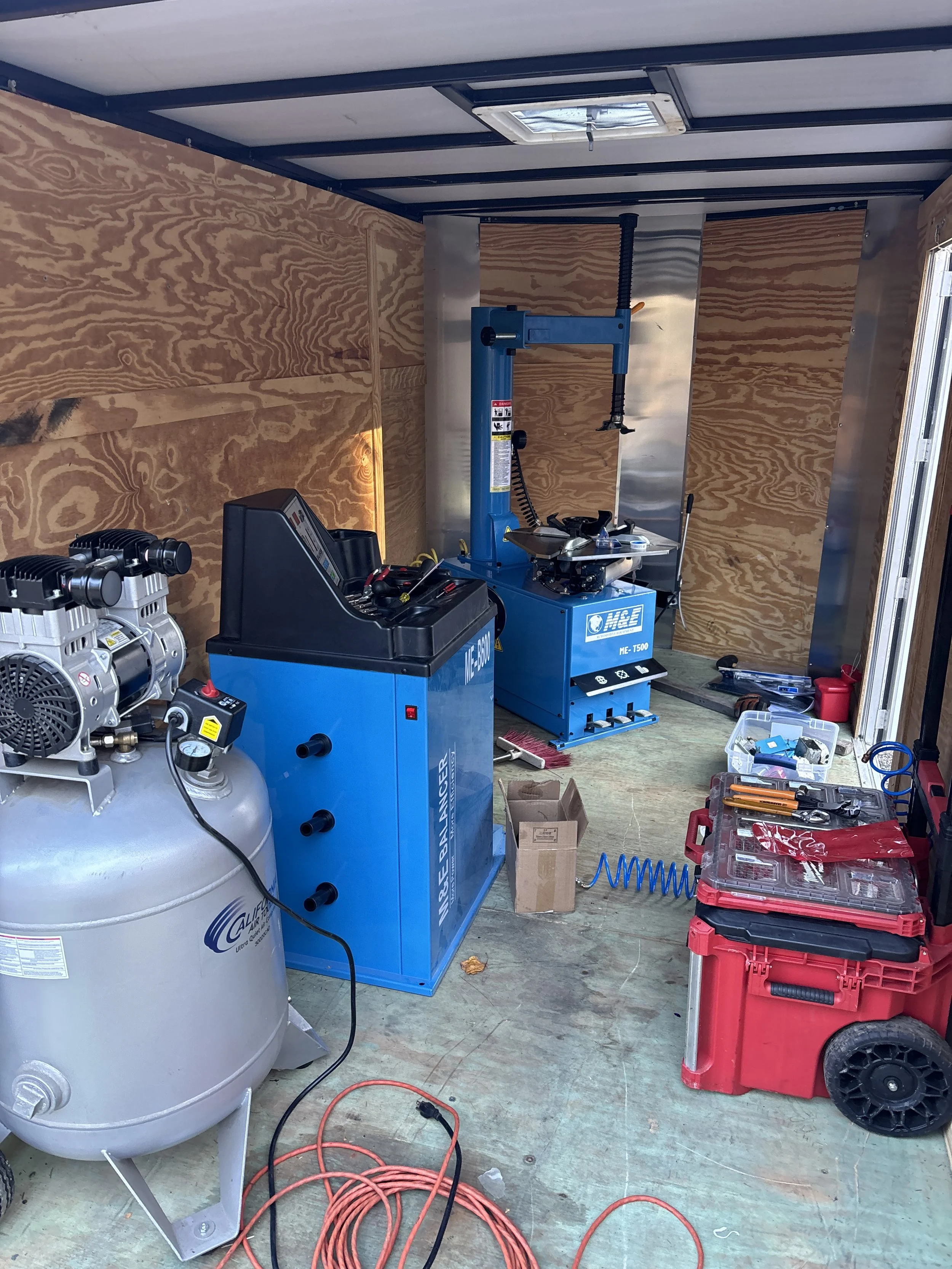 This image shows a workshop or garage with various tools and equipment, including an air compressor, a tire changer machine, a tool box with tools, and red storage bins. The walls are made of plywood, and there is a small vent or skylight in the ceiling.
