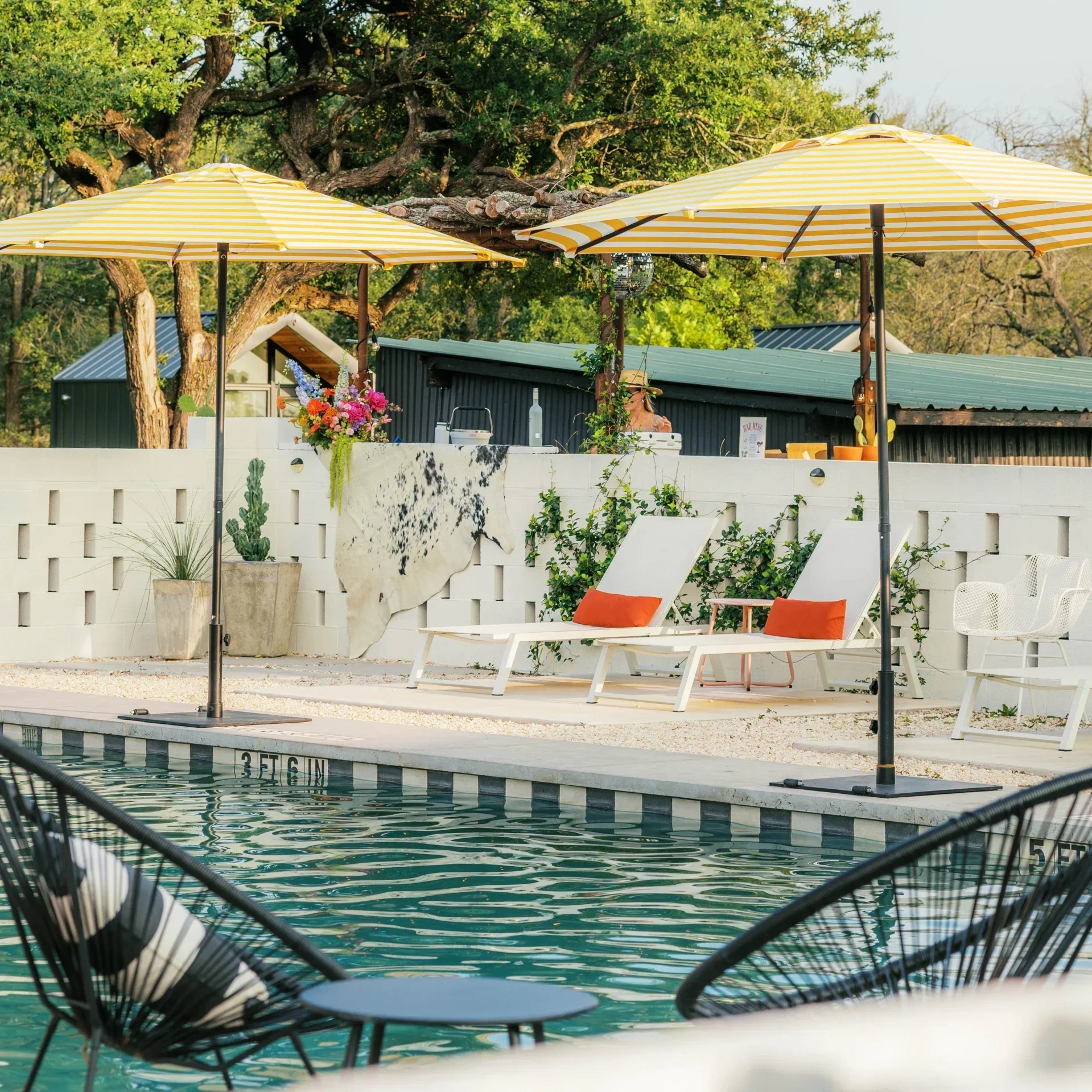 Poolside lounge chairs by a swimming pool with a black metal fence in the foreground, yellow umbrellas for shade, a person with a hat near a bar, lush green trees and a building with a metal roof in the background.