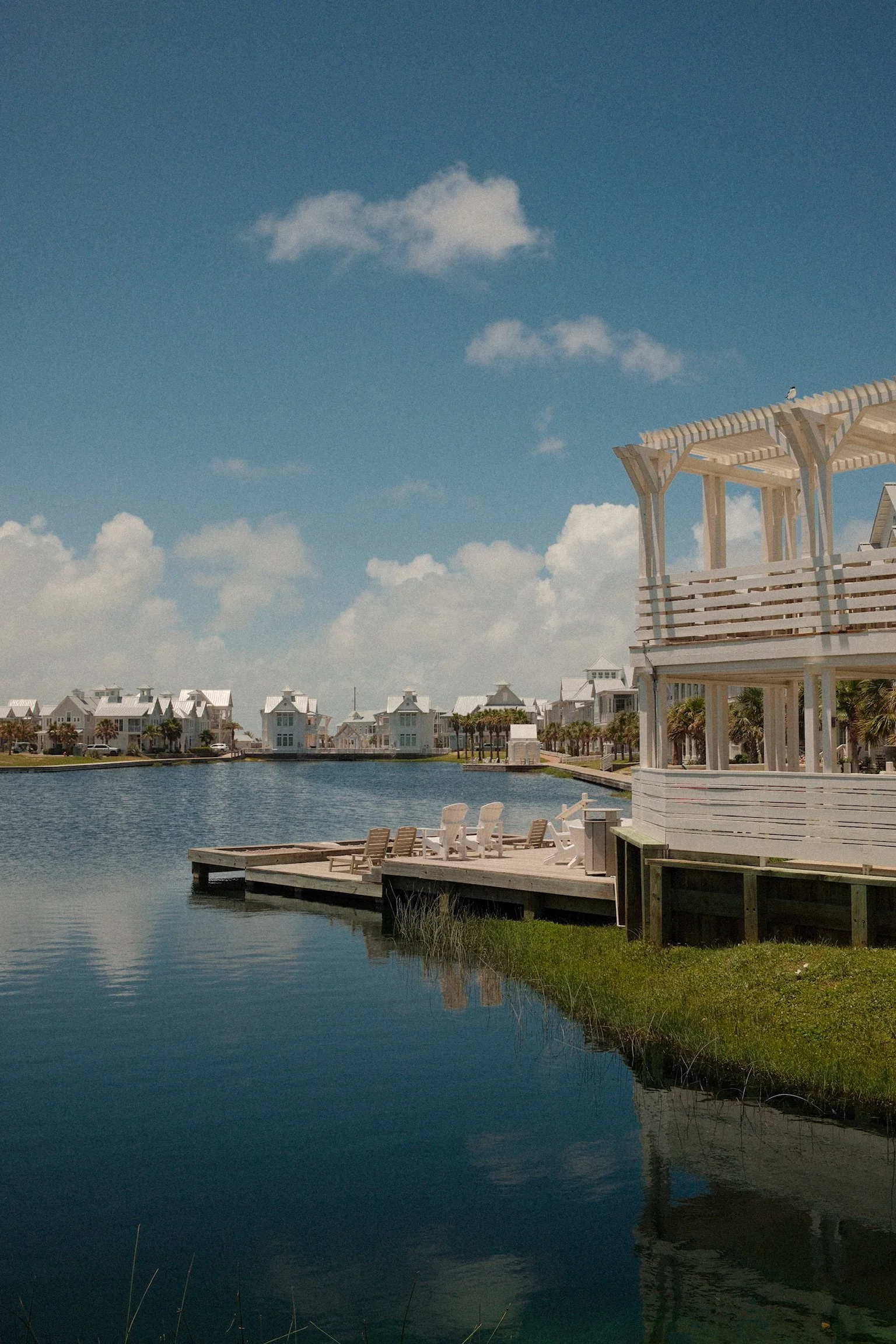 A waterfront scene with white buildings, a dock with lounge chairs, and a large white pergola on the right. The sky is blue with a few clouds, and the water reflects the structures.