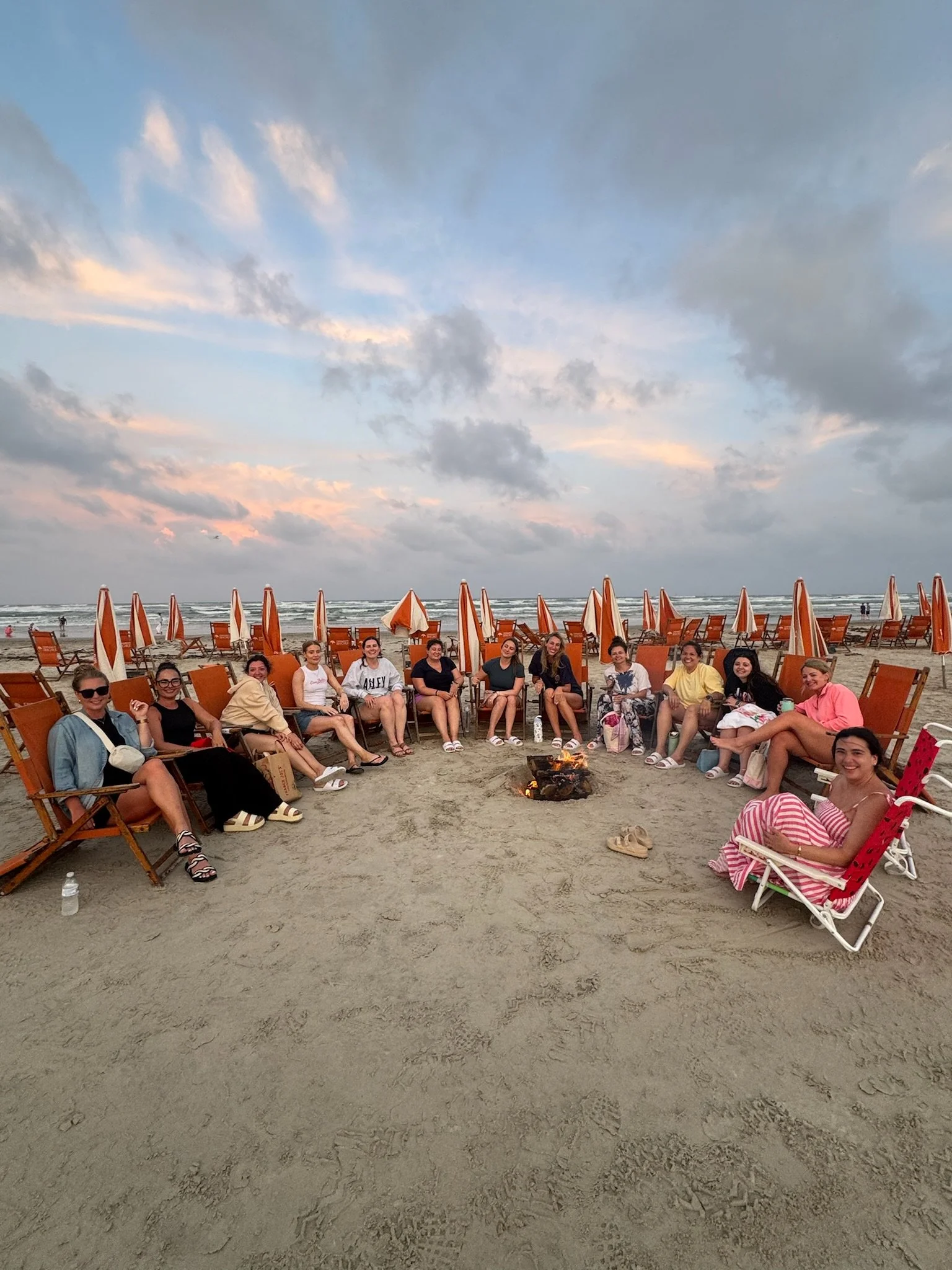 Group of women and children sitting around a small campfire on a sandy beach, with orange beach chairs and umbrellas, under a partly cloudy sky during sunset.