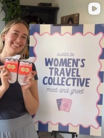 A woman holding two packs of snacks standing next to a sign that reads 'Austin TX Women's Travel Collective Meet and Greet'.