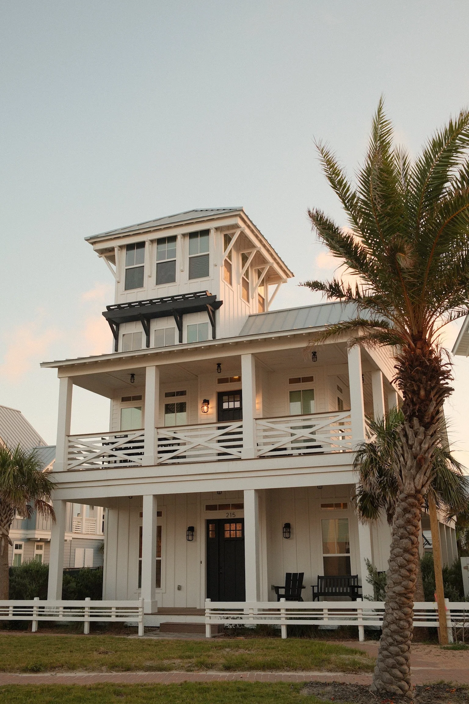 A white multi-story house with a tower, balcony, and porch, surrounded by palm trees, with a lawn and brick pathway in front, during sunset.
