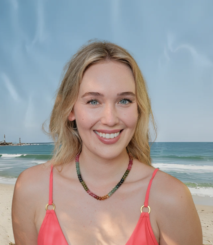 A young woman with blonde hair and blue eyes smiling at the camera at the beach.