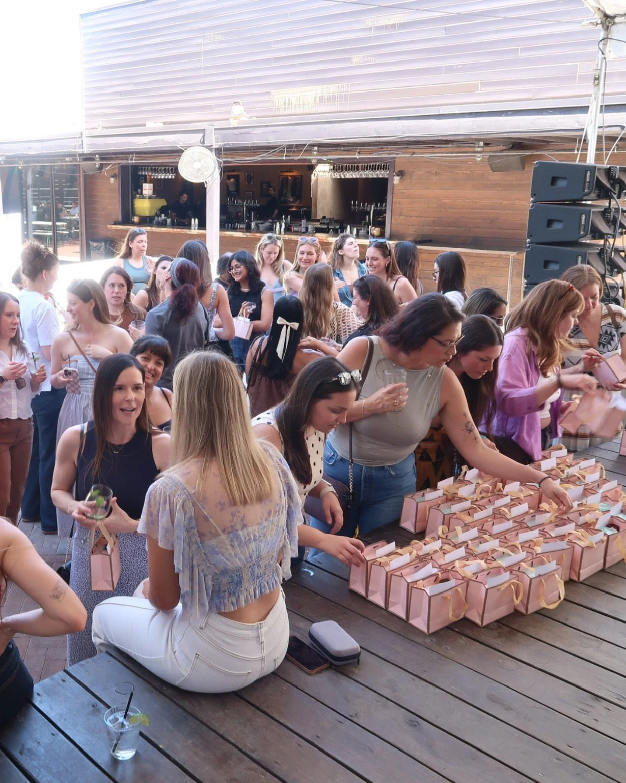 A group of women and girls gathered outdoors at a party or event, organizing pink gift bags on a wooden table. The setting appears to be a patio or outdoor venue with a bar in the background, and the crowd is engaged and chatting.