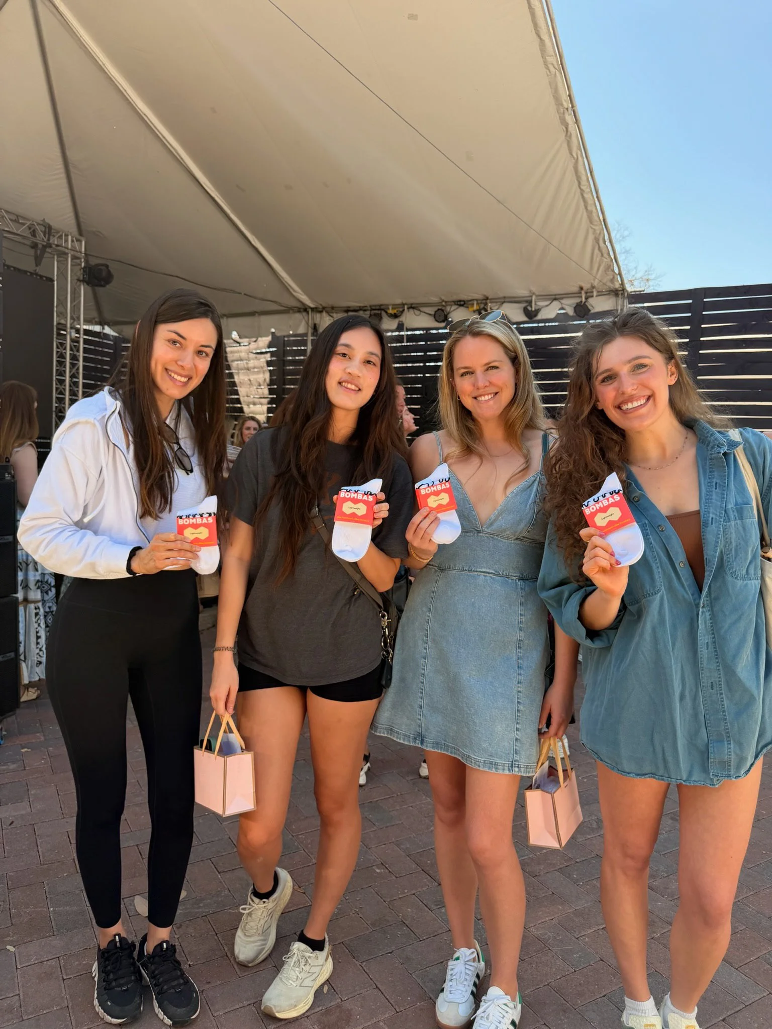 Four young women standing together outdoors in front of a tent at a daytime event, smiling at the camera and holding small gift bags and socks with red packaging that says "Bombas."