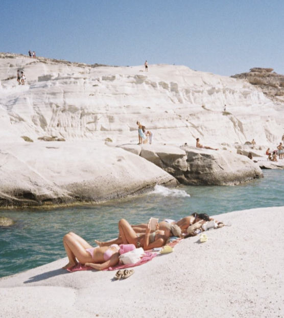 People sunbathing and relaxing on large rocks by the water near white cliffs under a clear blue sky.