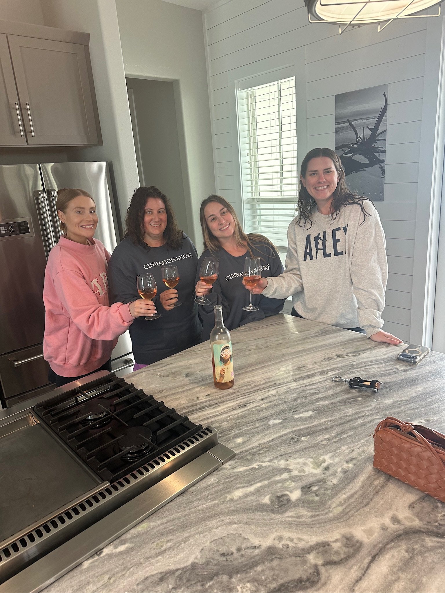 Four women smiling and holding glasses of wine inside a modern kitchen, with a marble countertop, a stainless steel refrigerator, and a black and white beach scene wall art.
