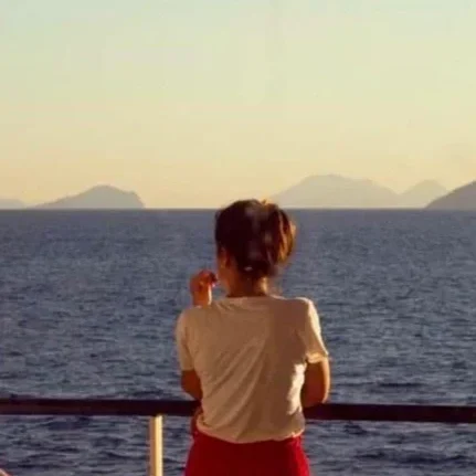A person standing on a boat or dock, facing away, looking out over the ocean with islands in the distance, during sunset or sunrise.