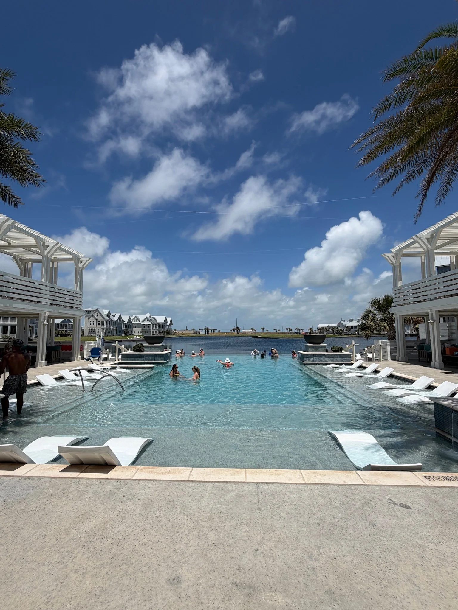 A swimming pool with people enjoying it, surrounded by white lounge chairs and white structures, overlooking a body of water with houses and palm trees in the background under a partly cloudy sky.