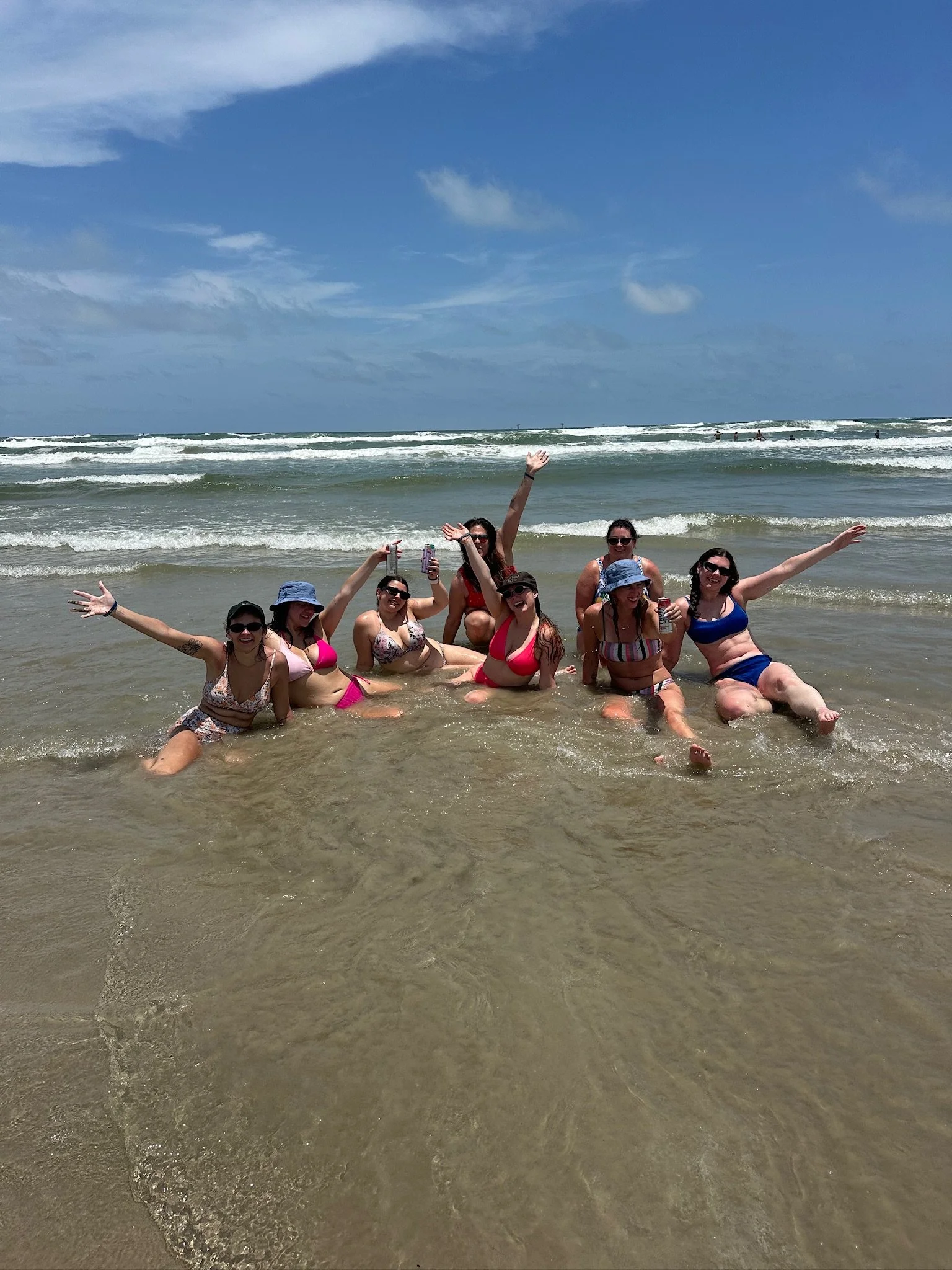 Group of women in swimsuits and hats enjoying and posing at the beach with ocean waves and blue sky in the background.