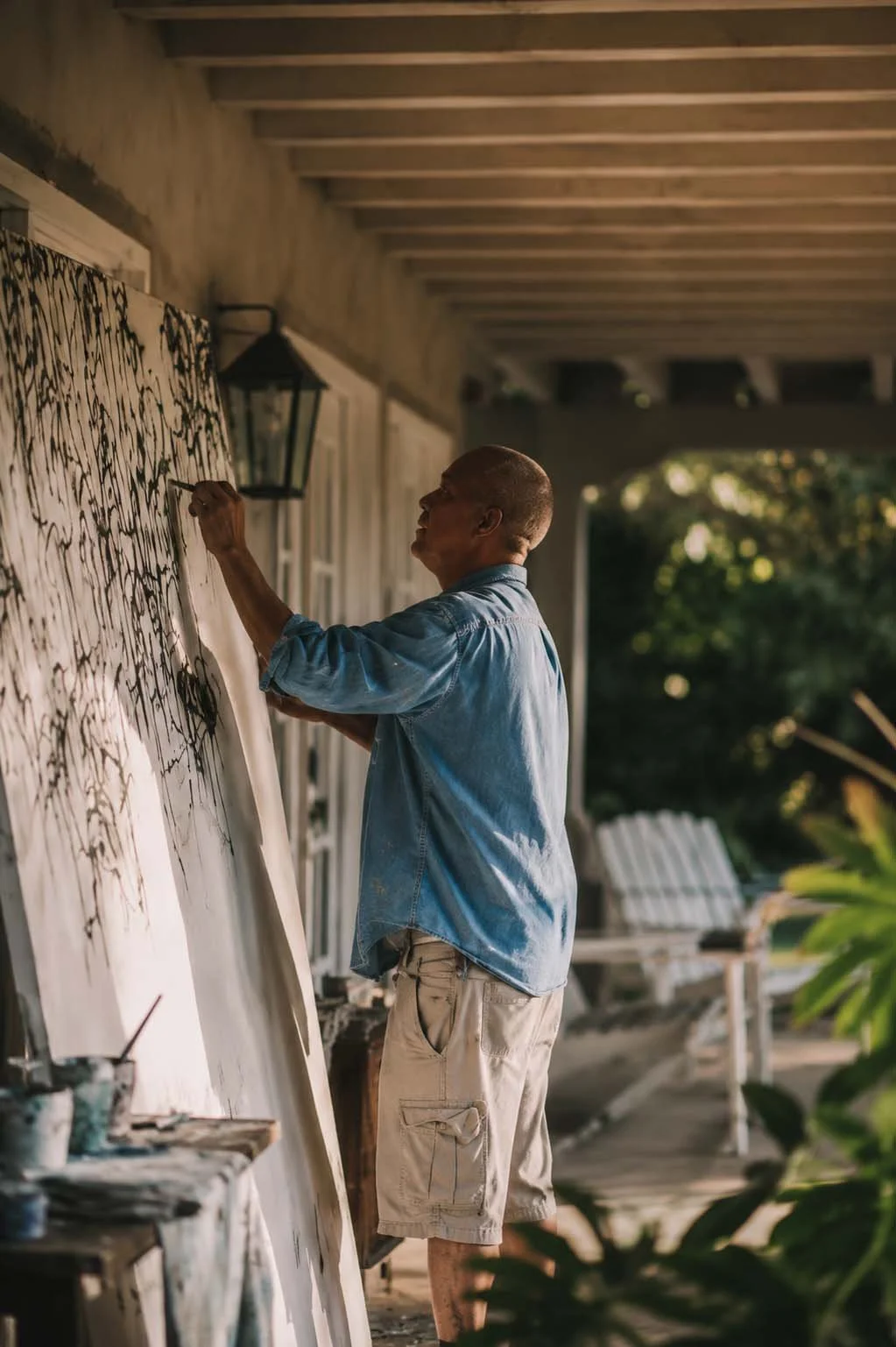 A man painting on a large canvas outdoors, under a porch, with trees and outdoor furniture visible in the background.