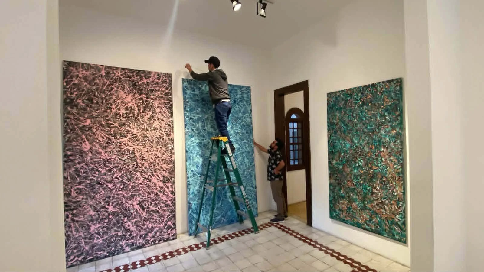 Two people in an art gallery hanging large, colorful abstract paintings on the wall. One person is standing on a ladder, attaching a painting, while the other is holding the ladder steady.