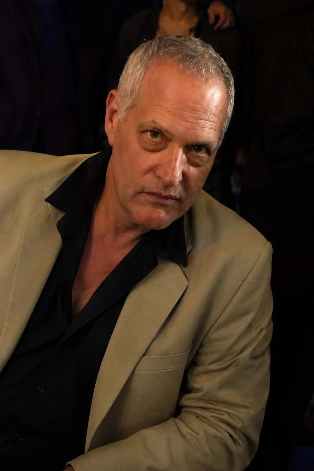 A middle-aged man with short gray hair wearing a beige blazer over a black shirt with an open collar, sitting indoors with a serious expression, and a blurred dark background with other people.