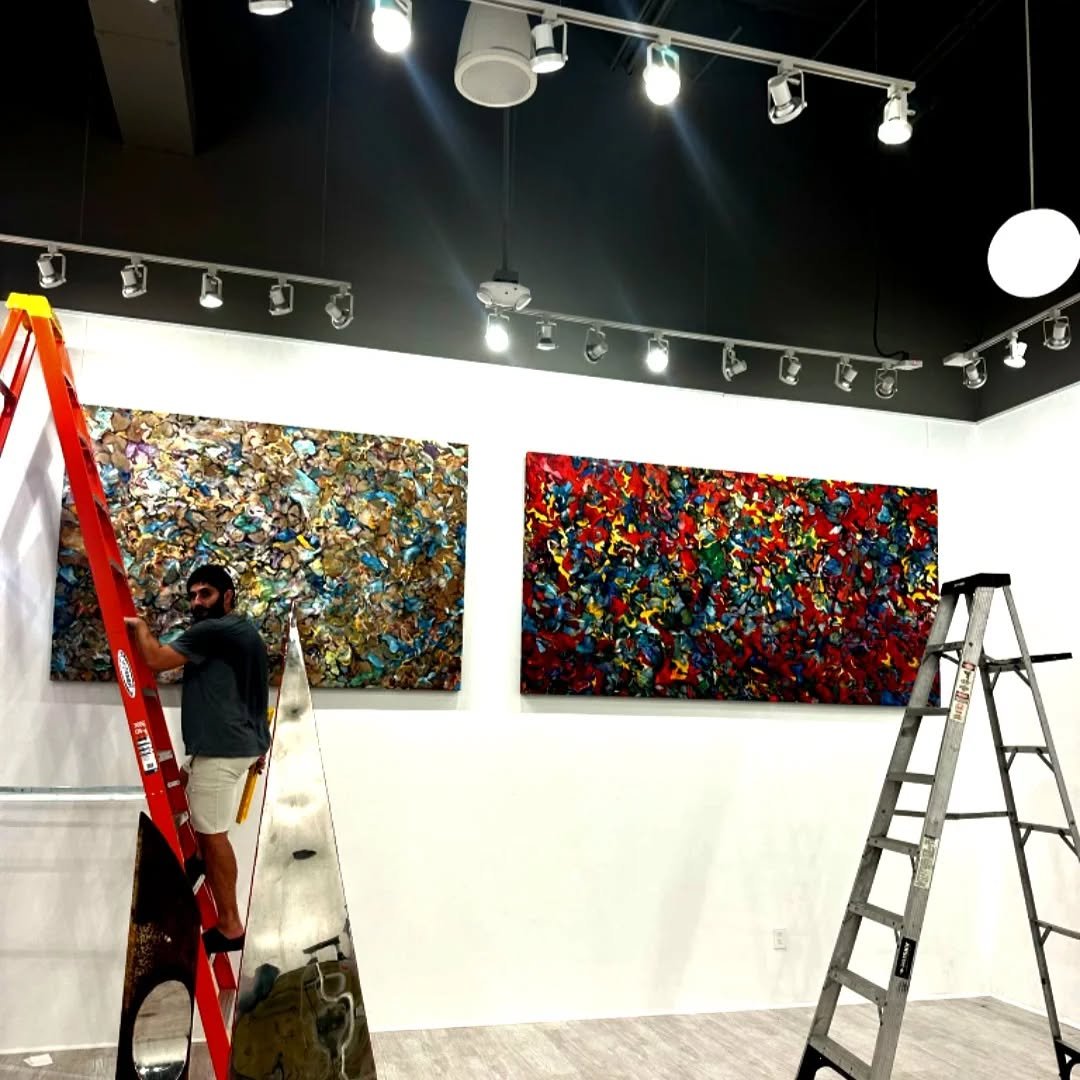 Art gallery with two large colorful abstract paintings on a white wall, two ladders, artificial lighting, and a person standing on a red ladder.