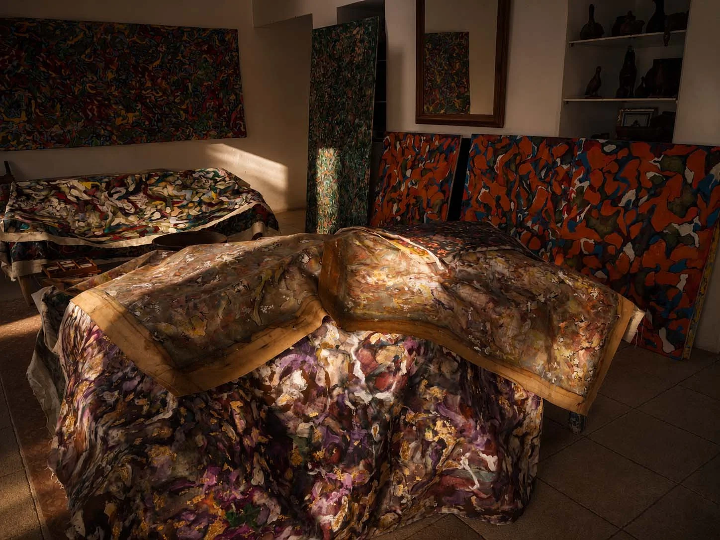 A room with several pieces of furniture covered in colorful, abstract-patterned fabrics. There is a bed, a chair, a mirror, and a shelf with small art pieces and a picture frame.