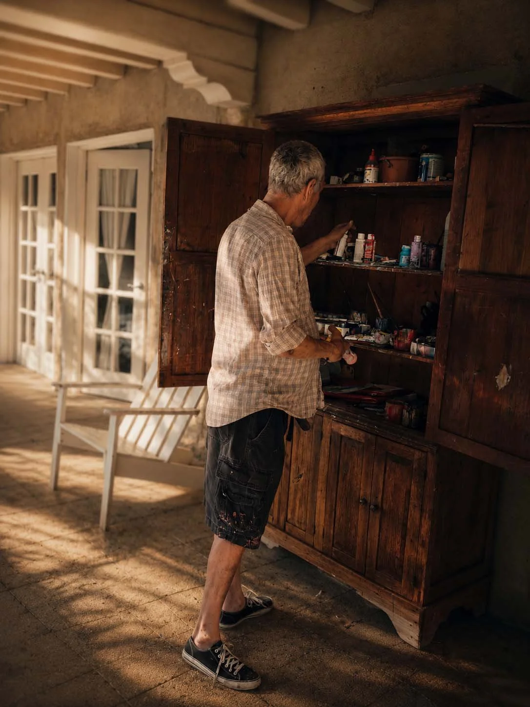 An elderly man with gray hair, wearing a plaid shirt, black shorts, and sneakers, is looking at and reaching into a wooden cabinet filled with various paint and art supplies. The setting appears to be a porch or outdoor patio with sunlight streaming in.