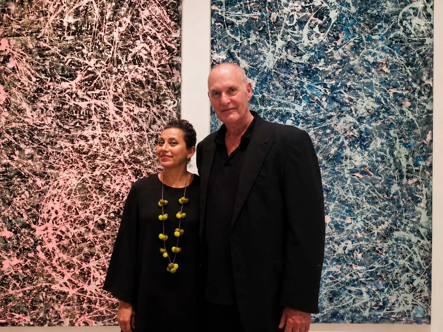 A woman and a man standing in front of two large, abstract paintings with intricate, colorful patterns.